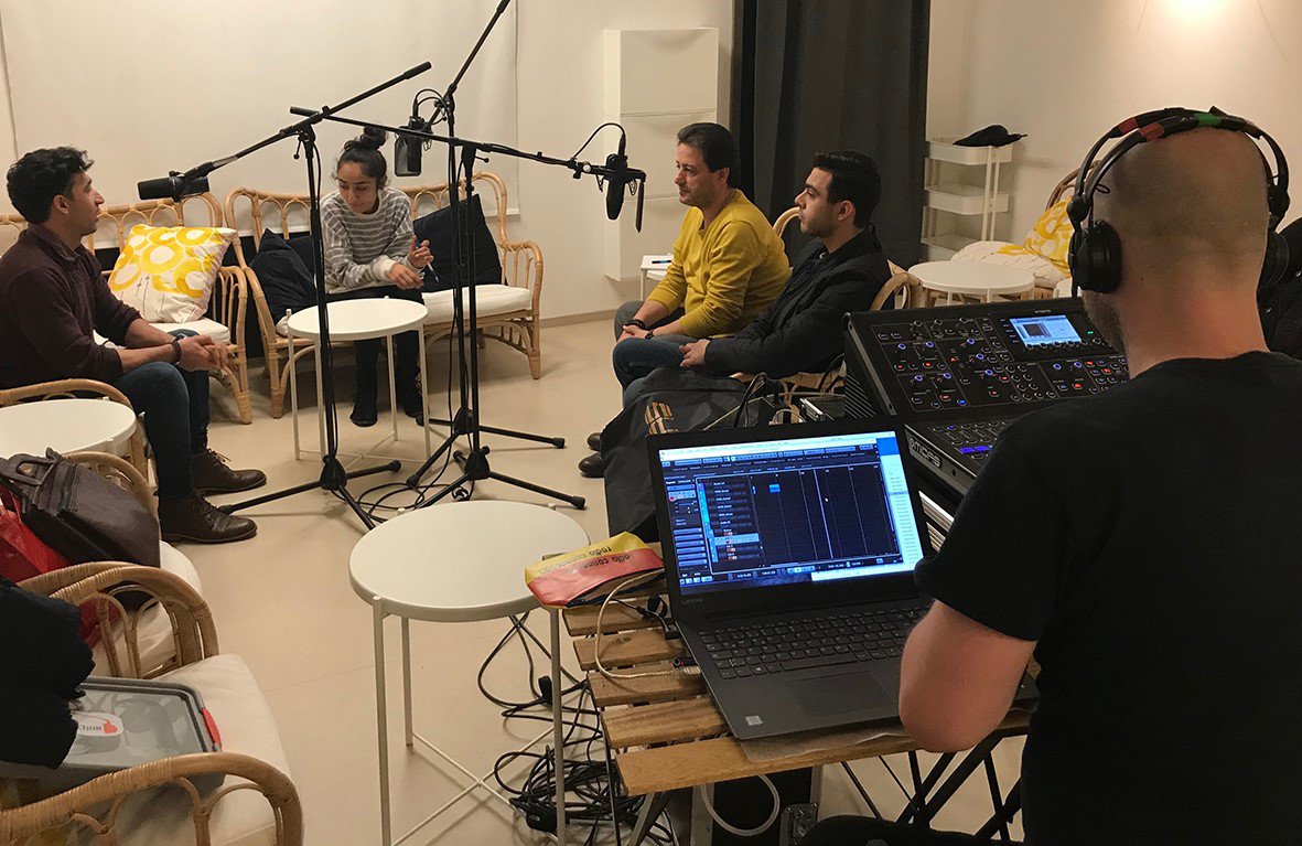 In einem Tonstudio machen 5 Personen eine Tonaufnahme. .  Im Vordergrund in der rechten Ecke des Bildes, sitzt ein Mensch mit einem Kopfhörer auf vor einem Computer und einem Mischpult.