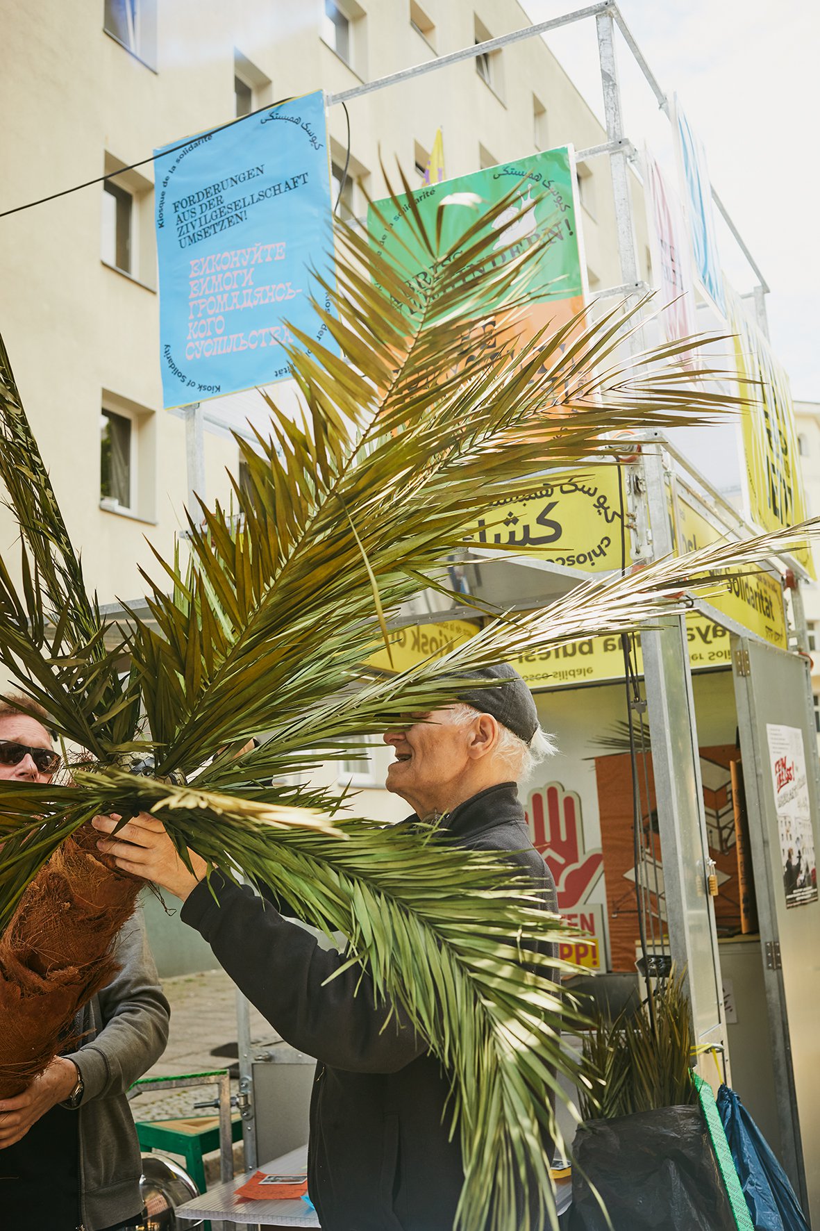 Im Vordergrund trägt ein junger Mann eine Palme. Er hält sie schräg, sodass ein älterer Herr an die Krone greifen kann. Im Hintergrund ist der Kiosk zu sehen.