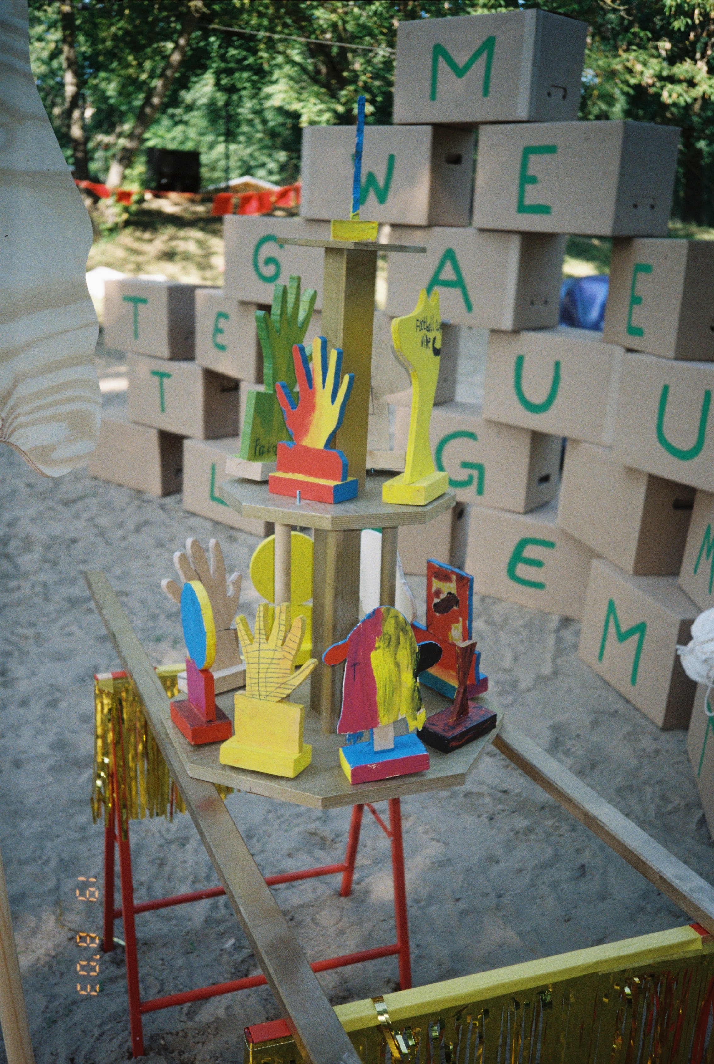 Eine Holzinstallation steht in einem Sandkasten in einem Park. Dahinter ist eine Wand aus Pappkartons auf die Buchstaben gemalt sind.