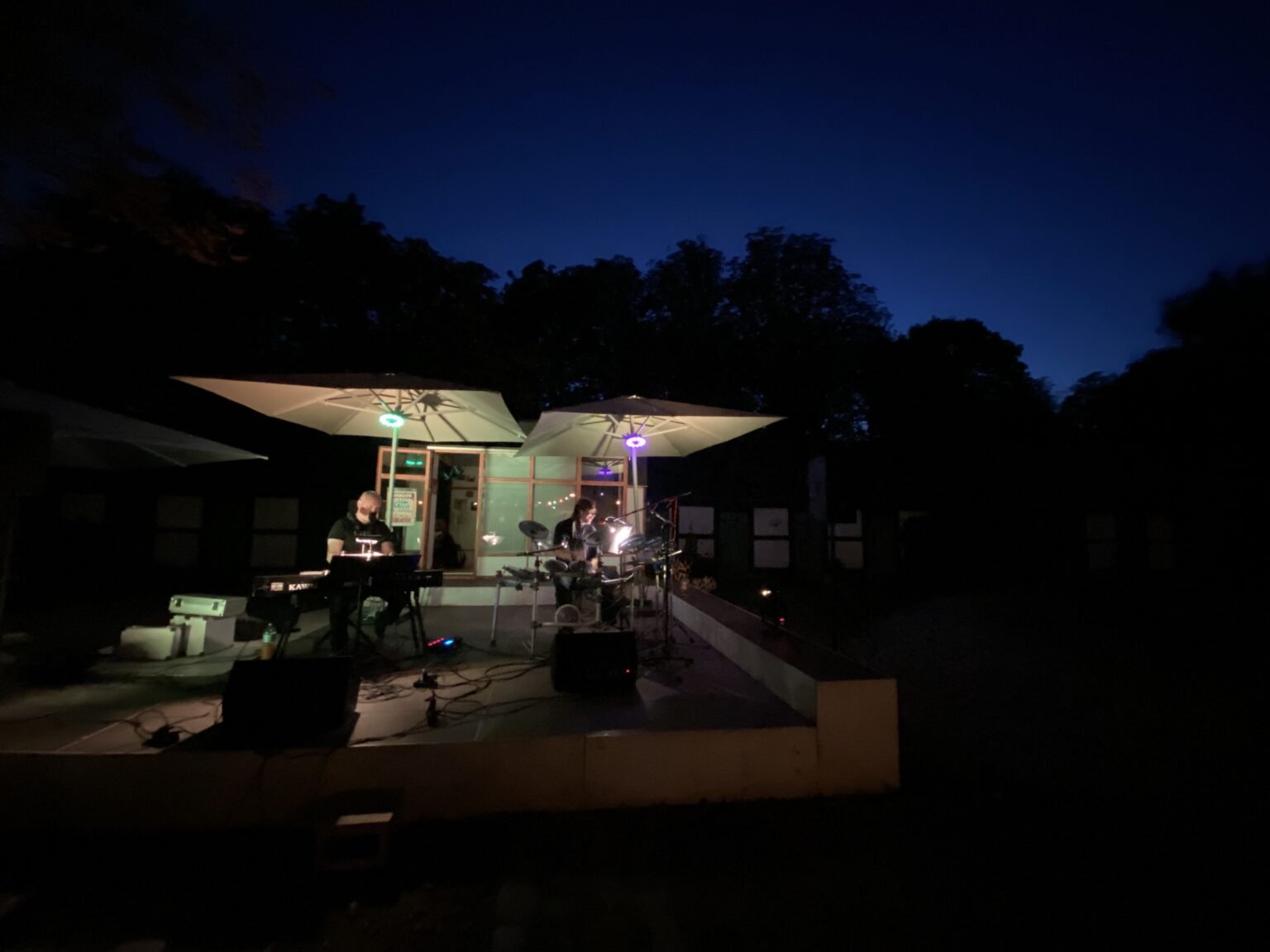 Open-Air-Konzert am sommerlichen Abend, es ist bereits dunkel. Auf der Bühne sitzt eine Person am Keyboard, daneben eine am Drumset. Beide Personen sitzen jeweils unter einem quadratischen hellem Schirm mit kleinen Scheinwerfern.