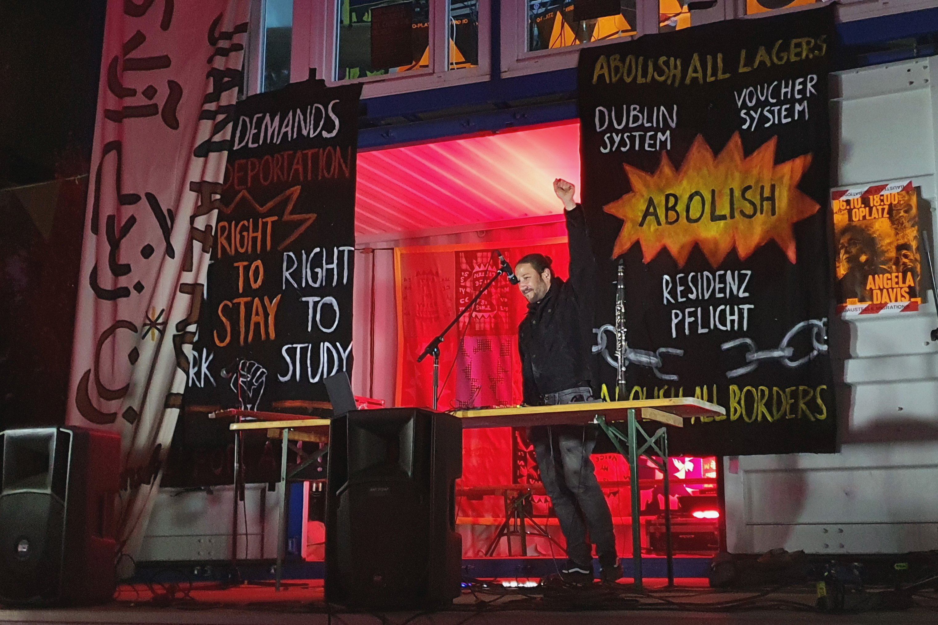 Ein Mann steht auf einer Bühne. Es ist Nacht. Er hält die Hand zur Faust geballt nach oben. Vor ihm steht ein Mikrophon. Hinter ihm sind Transparente an einem Holzgestell. Auf ihnen steht beispielsweise: Abolish all Lagers. Dublin System, Voucher System