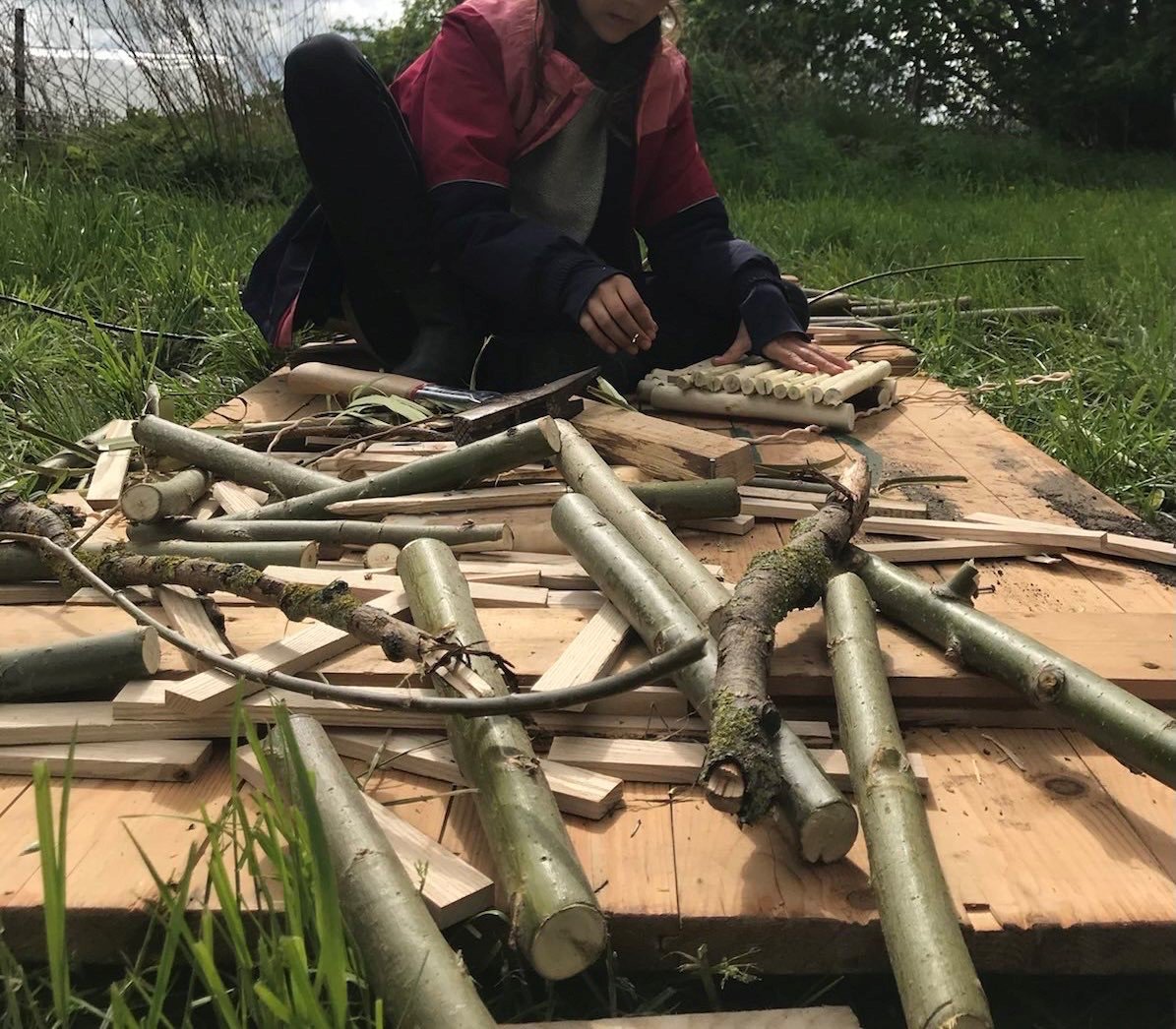 Auf dem Bild sind verschiedene Holzstücke und Äste zu sehen, die auf einer Holzplatte liegen. Im Hintergrund sind die Hände einer Person zu erkennen, die ein paar Holzstücke zusammenbaut.