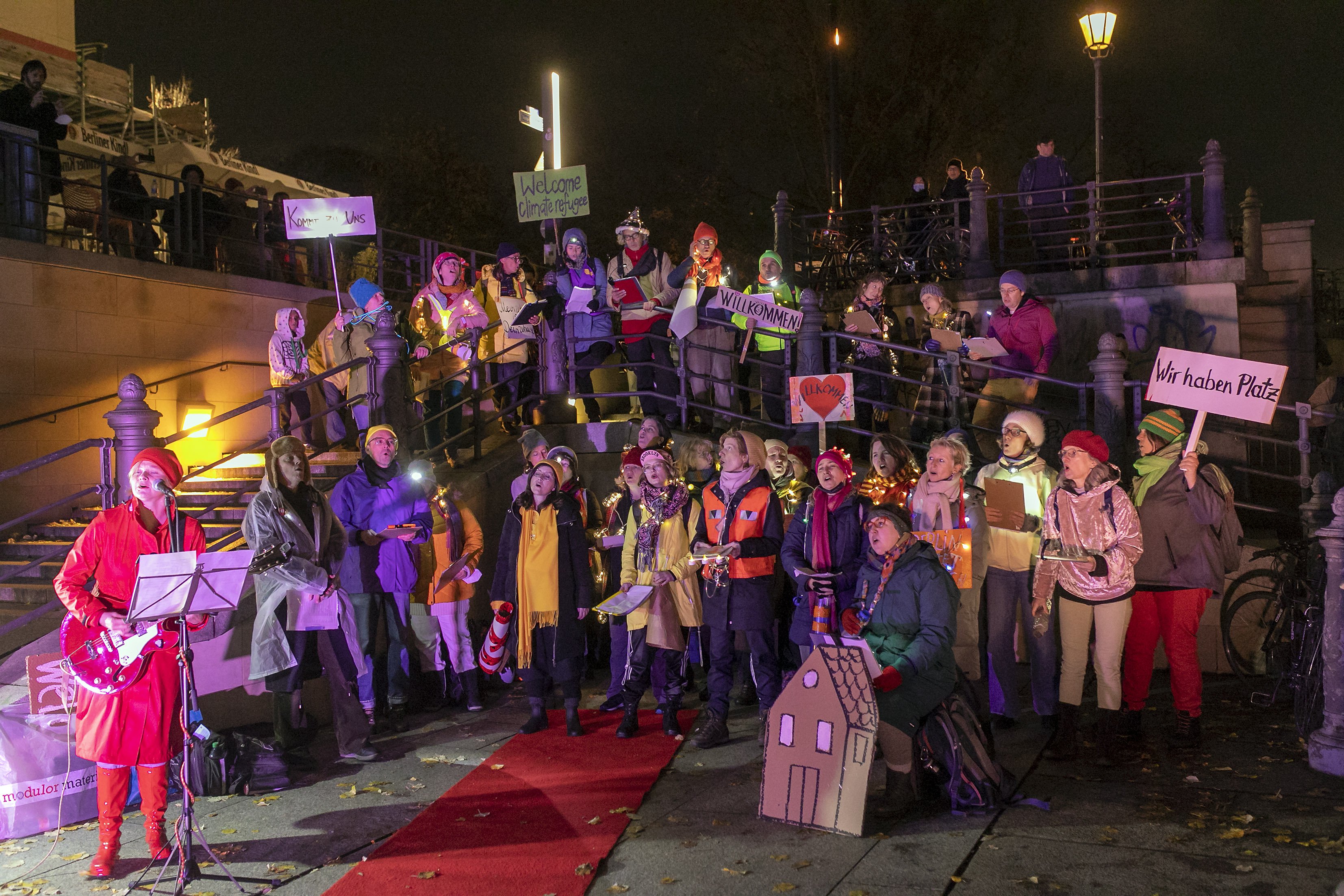 Der Chor der Statistik steht am Ufer der Spree und singt. Es ist abends und dunkel. Der Chor ist bunt beleuchtet. Mehrere Personen halten Plakate hoch. Auf einem steht "wir haben Platz".