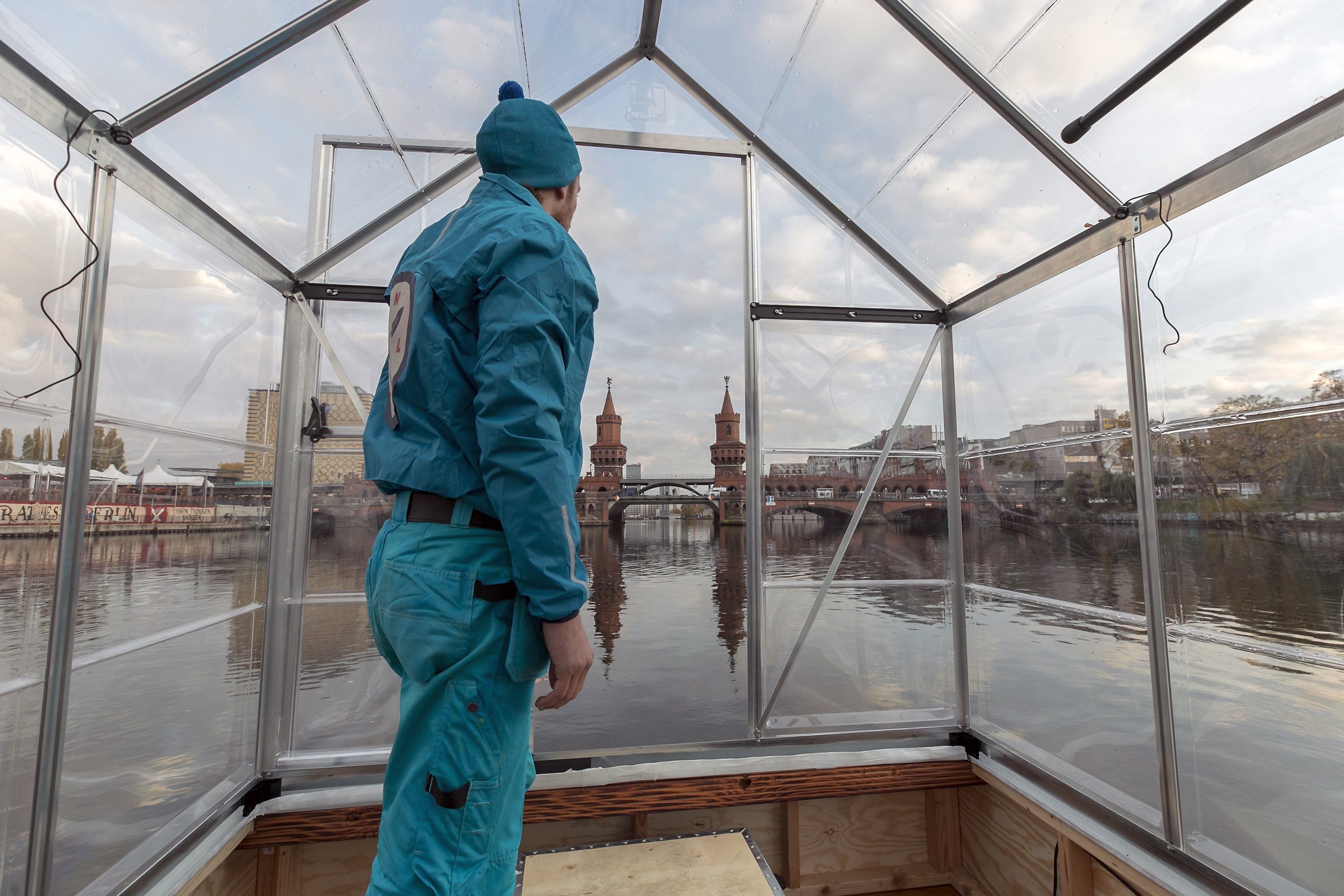 Eine Person steht aufrecht auf einem selbstgebauten Floß aus Plexiglas-Wänden. Das Floß ist auf der Spree. Die Person im türkisenen Outfit schaut von der Kamera weg Richtung Oberbaumbrücke in Kreuzberg.