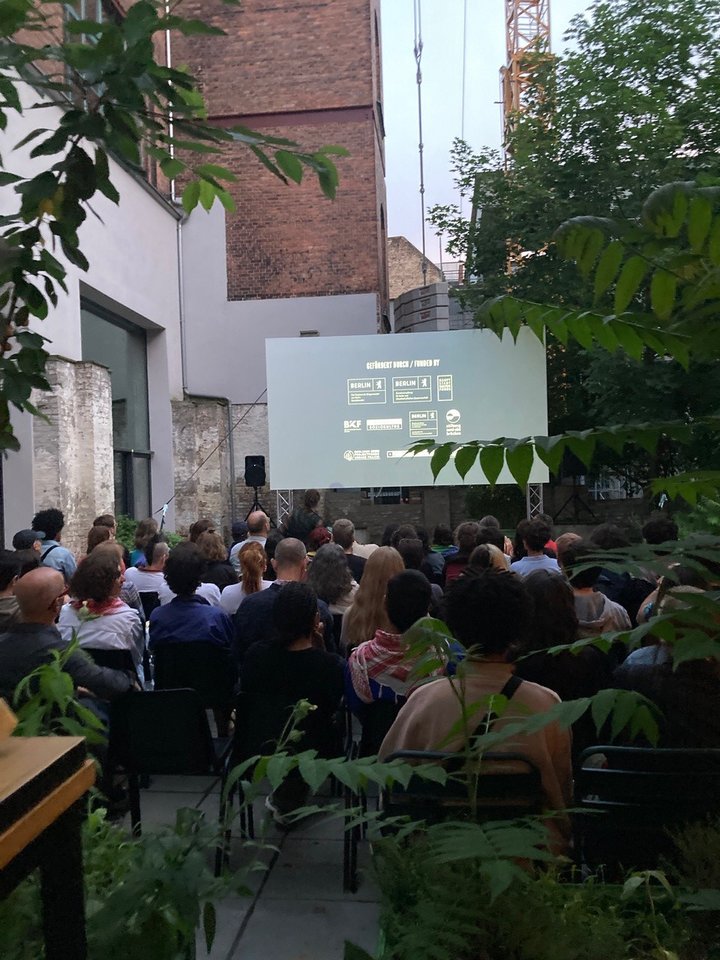 Ein Foto des Open Air Kino Bereichs des Kinos. Stuhlreihen sind aufgestellt in einem kleinen Hof. Ganz hinten ist eine Leinwand aufgehängt auf der ein Film läuft.