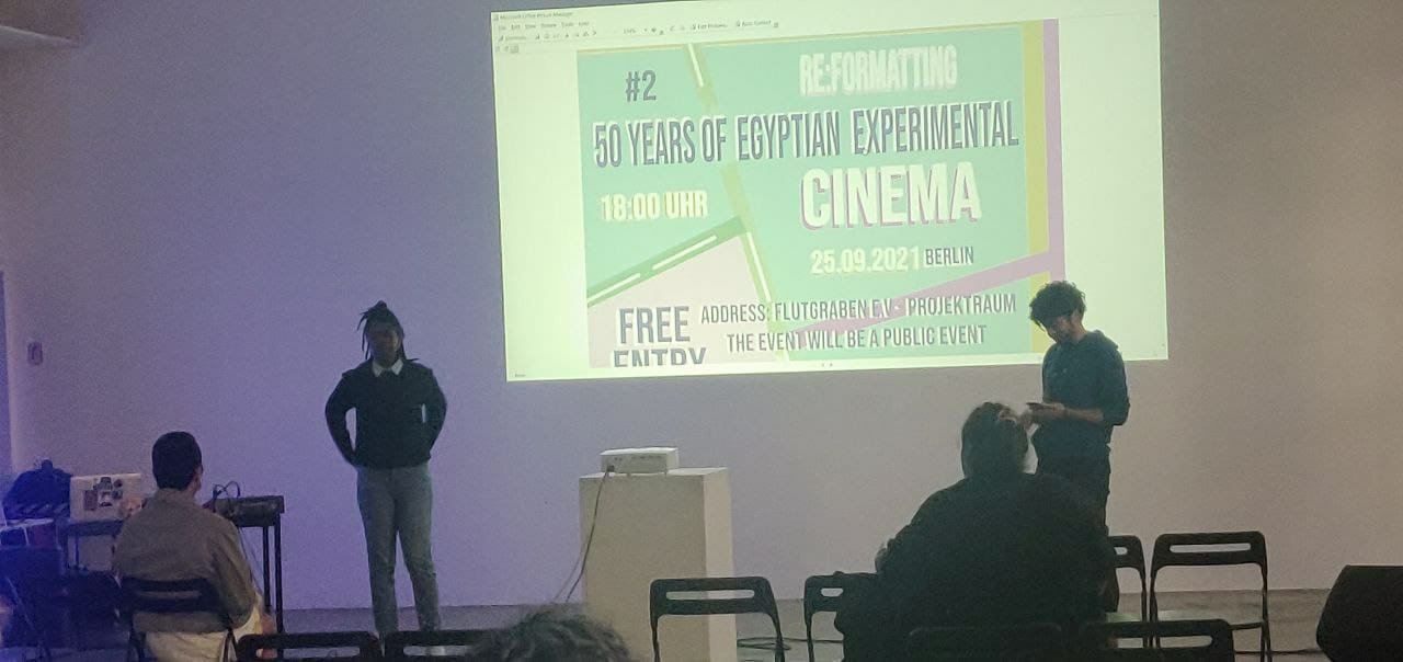 An die Wand ist der Flyer der Veranstaltung „Re:formation 50 Years of Egyptian experimental Cinema“ projiziert. Zwei Personen stehen rechts und links vom projizierten Bild dem Publikum zugewandt.