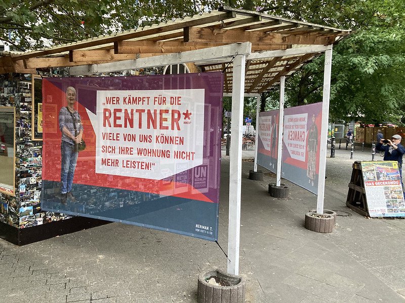 Eine Rentnerin ist auf einem großen Transpi abgebildet, das am Gecekondu hängt. Auf dem Transpi steht: Wer kämpft für die Renter*innen? Viele von uns können sich ihre Wohnung nicht mehr leisten. Darunter ist ihr Name, Neriman T., geschrieben.