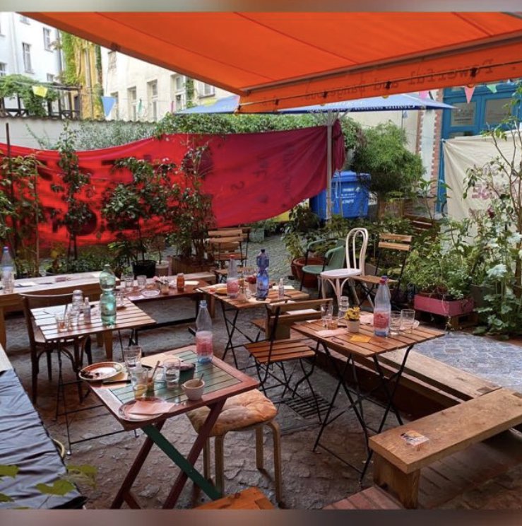 Der Hinterhof der Oya-Bar ist mit vielen Café-Tischen und Stühlen bestückt. Auf allen 6 Tischen stehen angebrochene Getränke. Eine orangene Markise ist über den Hof ausgefahren, es ist Sommer.