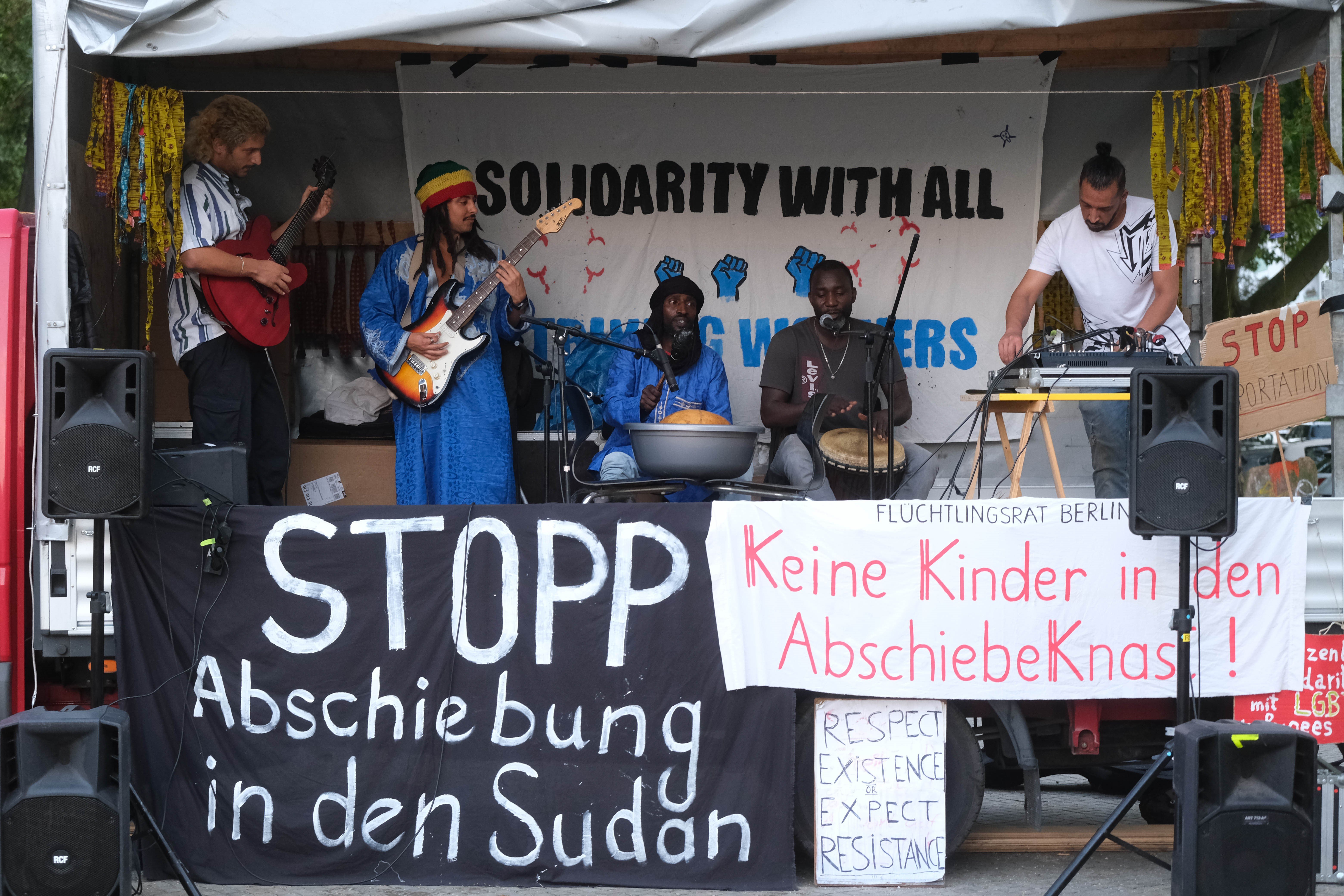 Eine Band spielt auf dem Hänger eines Autos. Es hängen Stoffbanner auf dem Hänger auf denen stehen: Stopp die Abschiebungen in den Sudan.