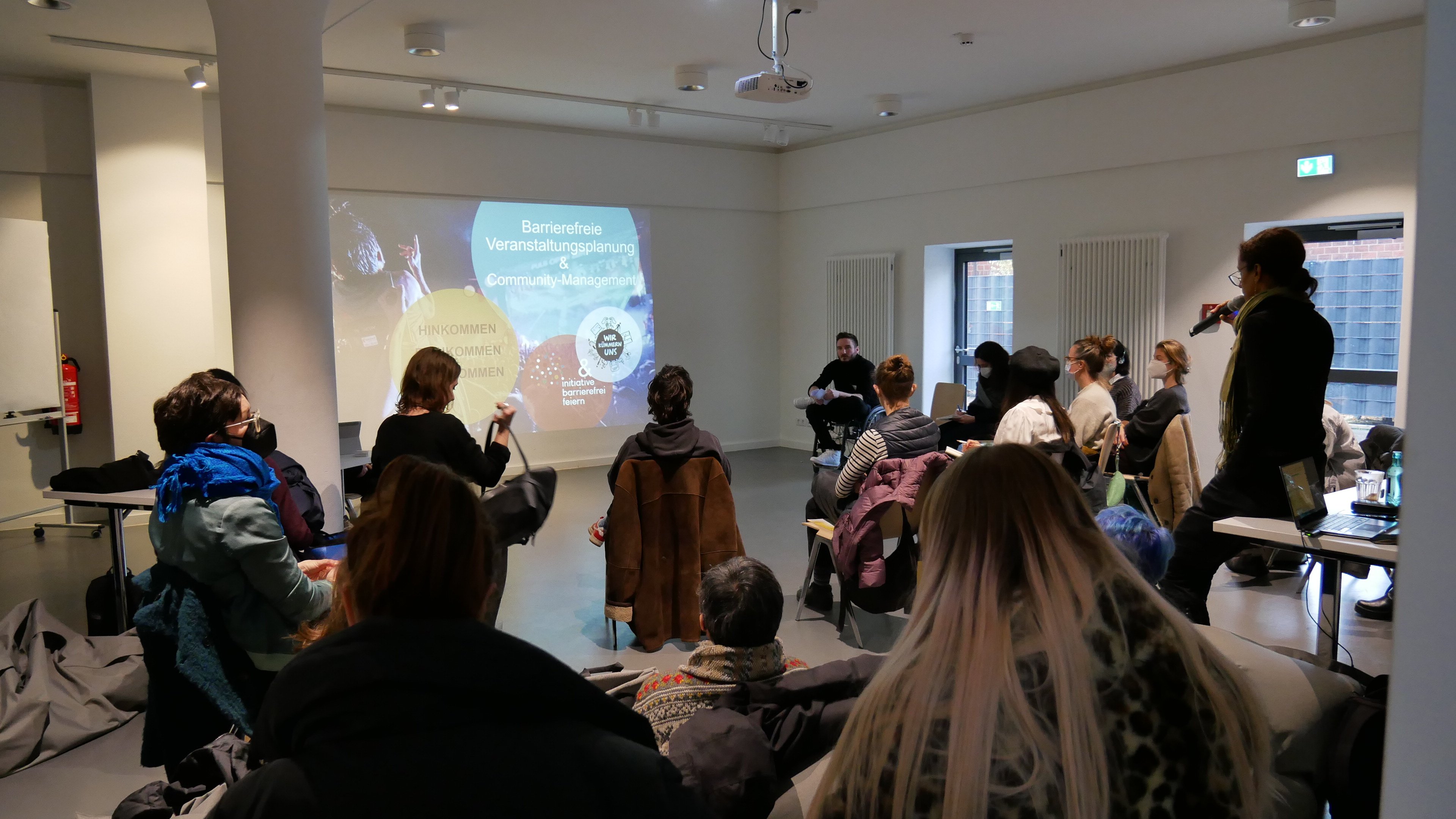 Ein Workshop der Initiative Barrierefrei feiern. Eine kleine Gruppe sitzt im Halbkreis. Im Hintergrund ist ein Bild an die Wand projeziert auf dem steht: Barrierefreie Veranstaltungsplanung und Community Management.