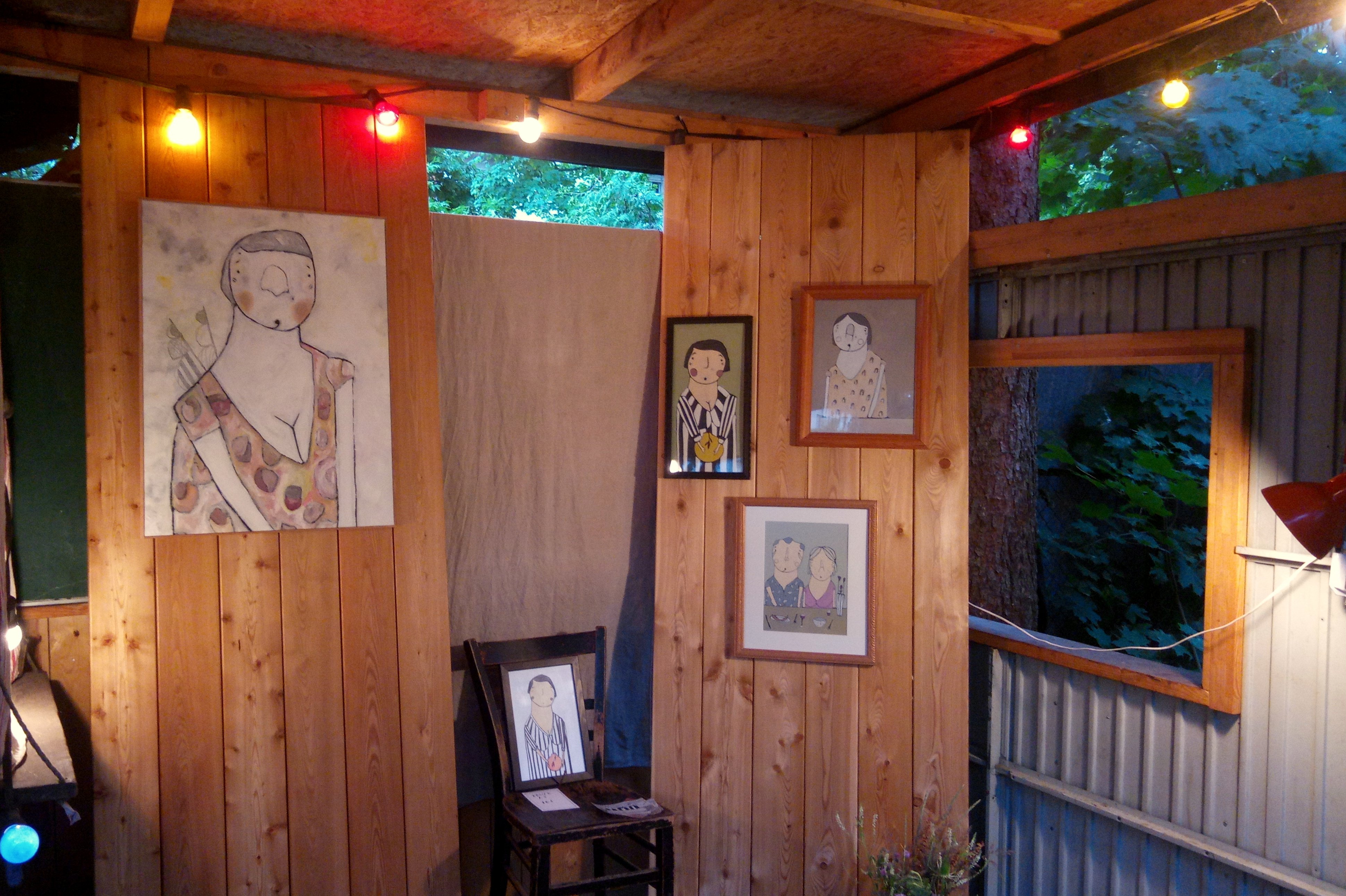 Im Innenraum des kleinen Holzhauses hängen gezeichnete Portraits von V.D. Blokk. Sie sind skizzenhaft coloriert. Sie hängen in unterschiedlicher Anordnung und Größe. Ein kleineres gerahmtes Bild steht in der Bildmitte auf einem Holzstuhl.