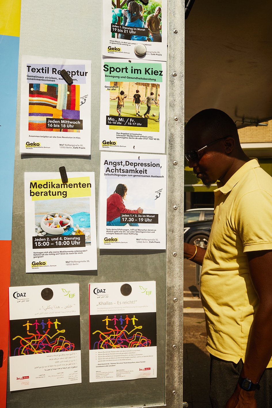 Die Seitenwand des Kiosk. Auf ihr kleben verschiedene Zettel mit Angeboten, wie Sport im Kiez, Textil-Reparatur, Medikamentenberatung und Angst, Depressionen, Achtsamkeit. Rechts steht ein Mann und blickt auf die Hinterseite des Kiosk.