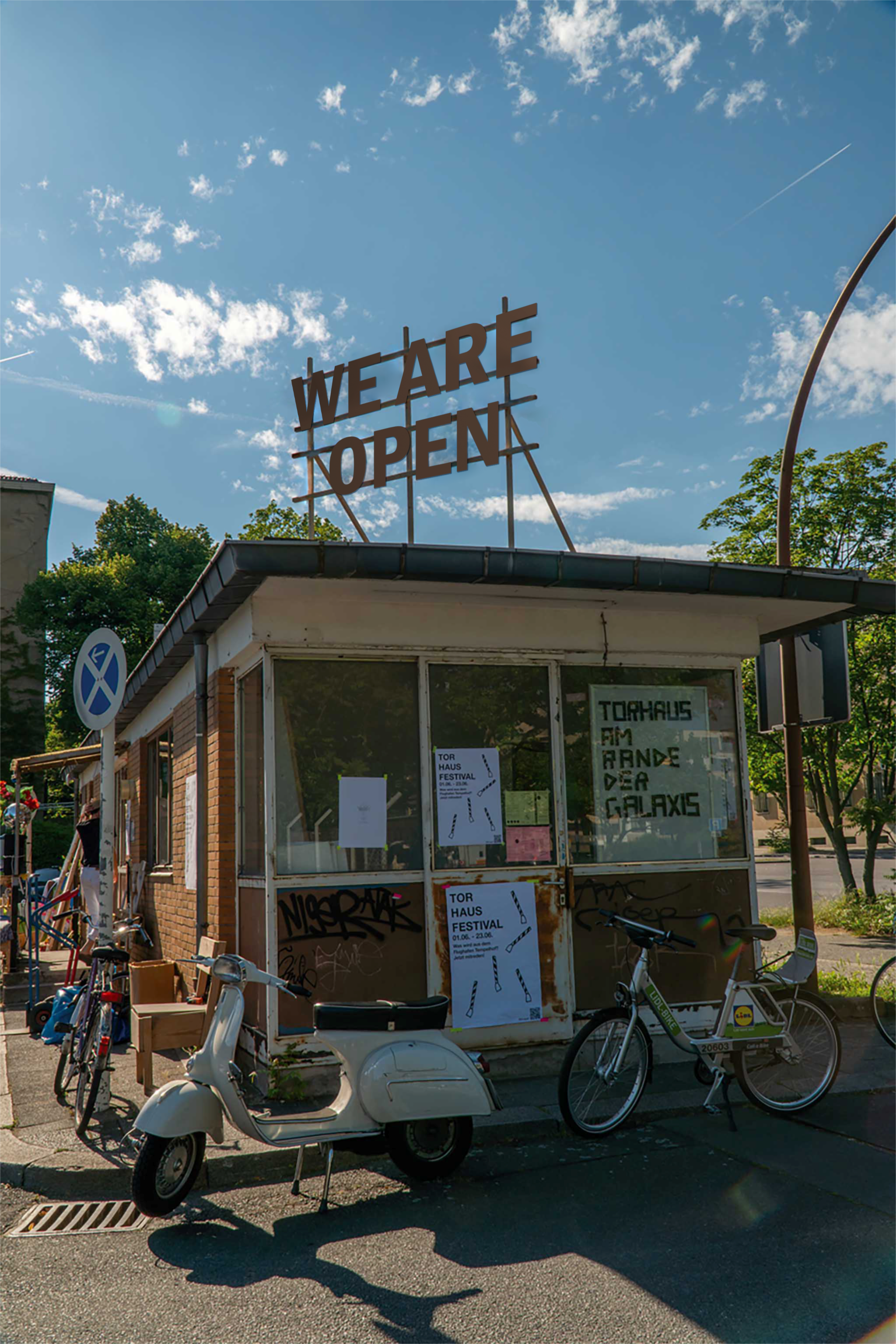 Eine kleine Hütte, in der Mitte eines großen Bürgersteigs zwischen zwei Straßen. In den Fenstern hängen Plakate. Auf dem Haus ist ein Schriftzug an zwei Stäben befestigt. Dort steht: #We are Open