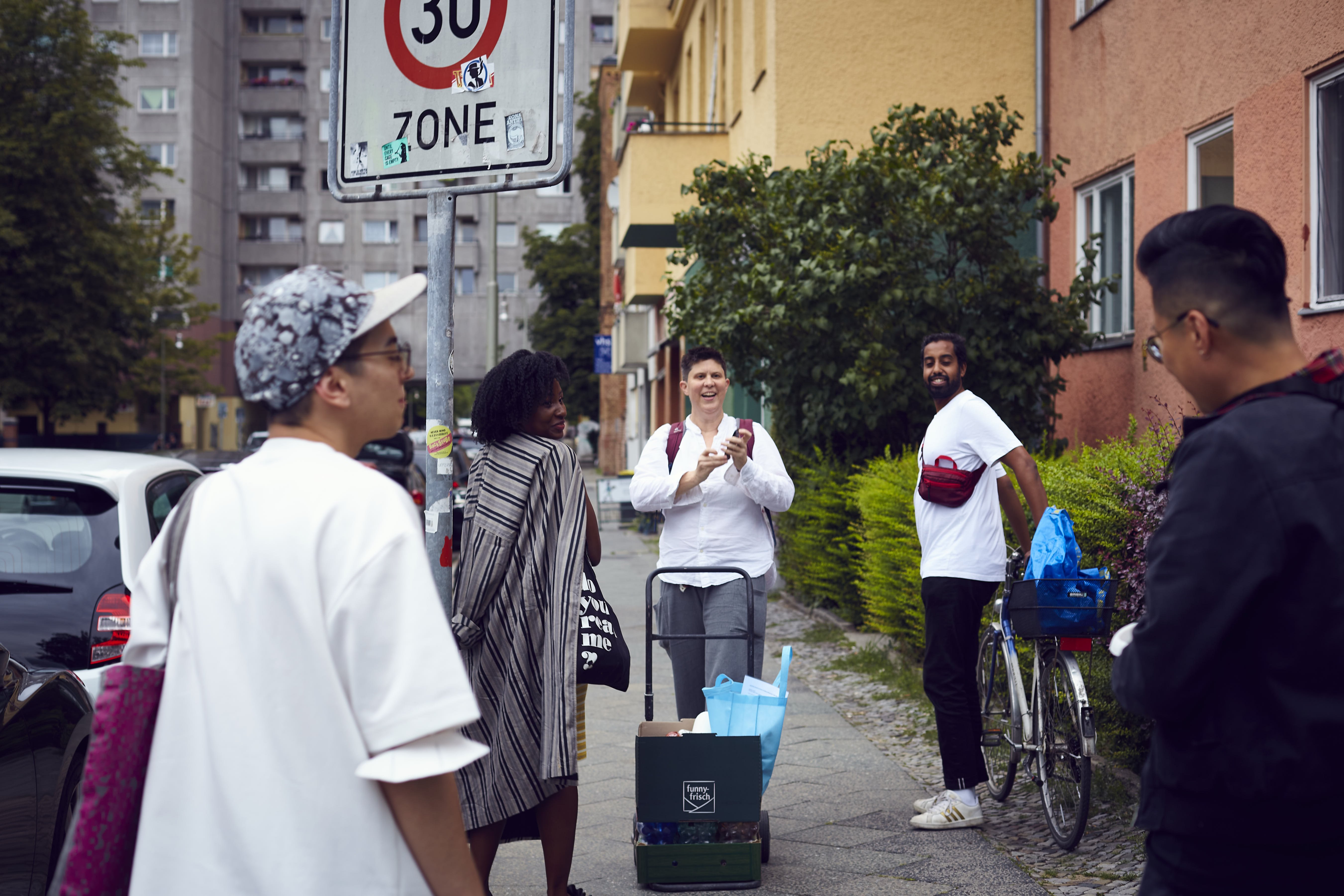 5 Menschen stehen auf dem Bürgersteig in der Wassertorstraße in Kreuzberg. Zwei Menschen im Vordergrund verschwommen, sind die drei Menschen im Zentrum des Bildes scharf zu erkennen. Im Hintergrund sieht man Mehrfamilienhäuser, Büsche und einen Baum.