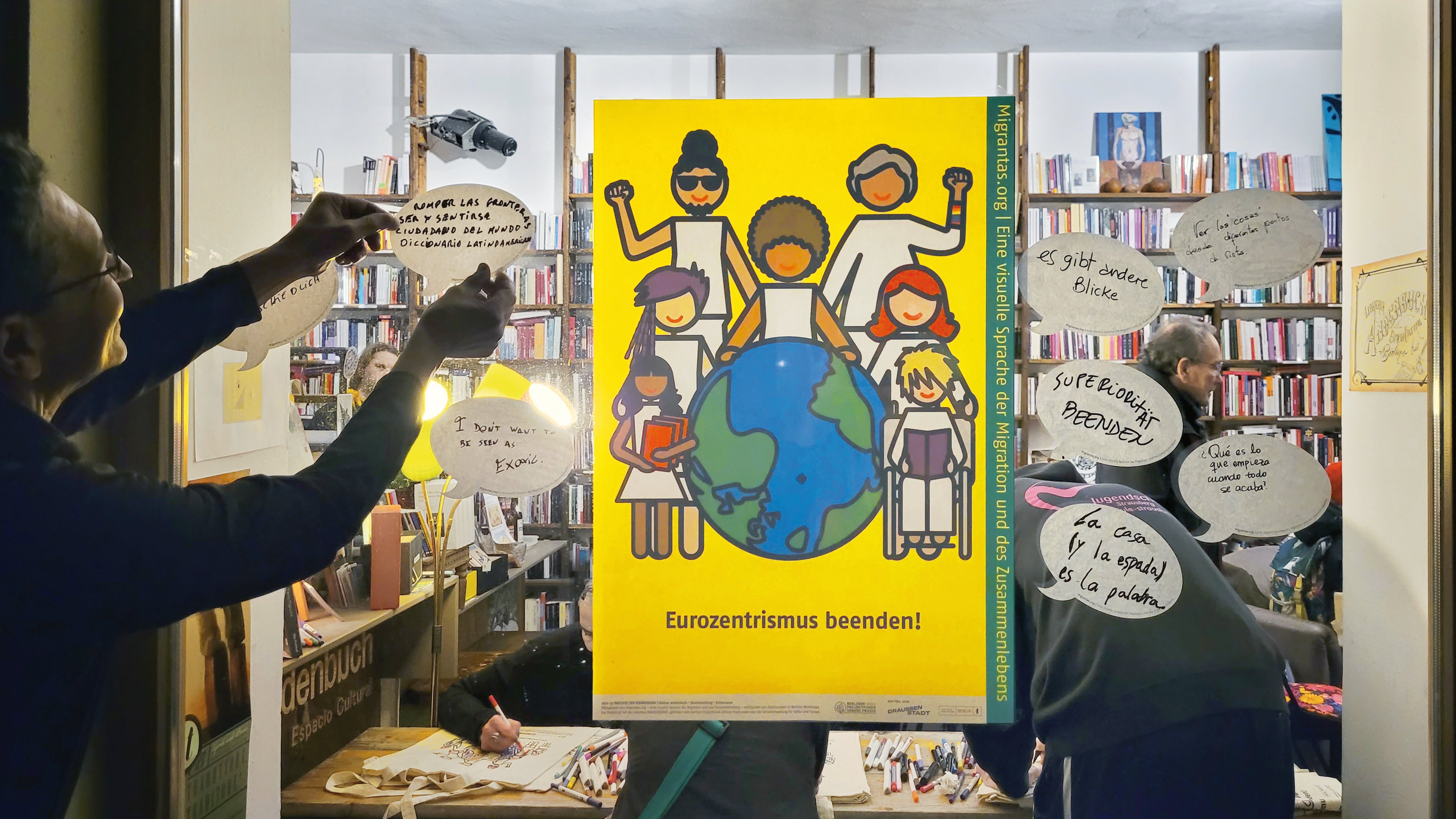 Ein Plakat hängt in einem Fenster. Auf dem Plakat steht Eurozentrismus beenden! Darüber sind gemalte Figuren die um einen Globus aufgestellt sind. Eine Person hängt links neben dem Plakat eine Karte auf, auf der etwas geschrieben steht.