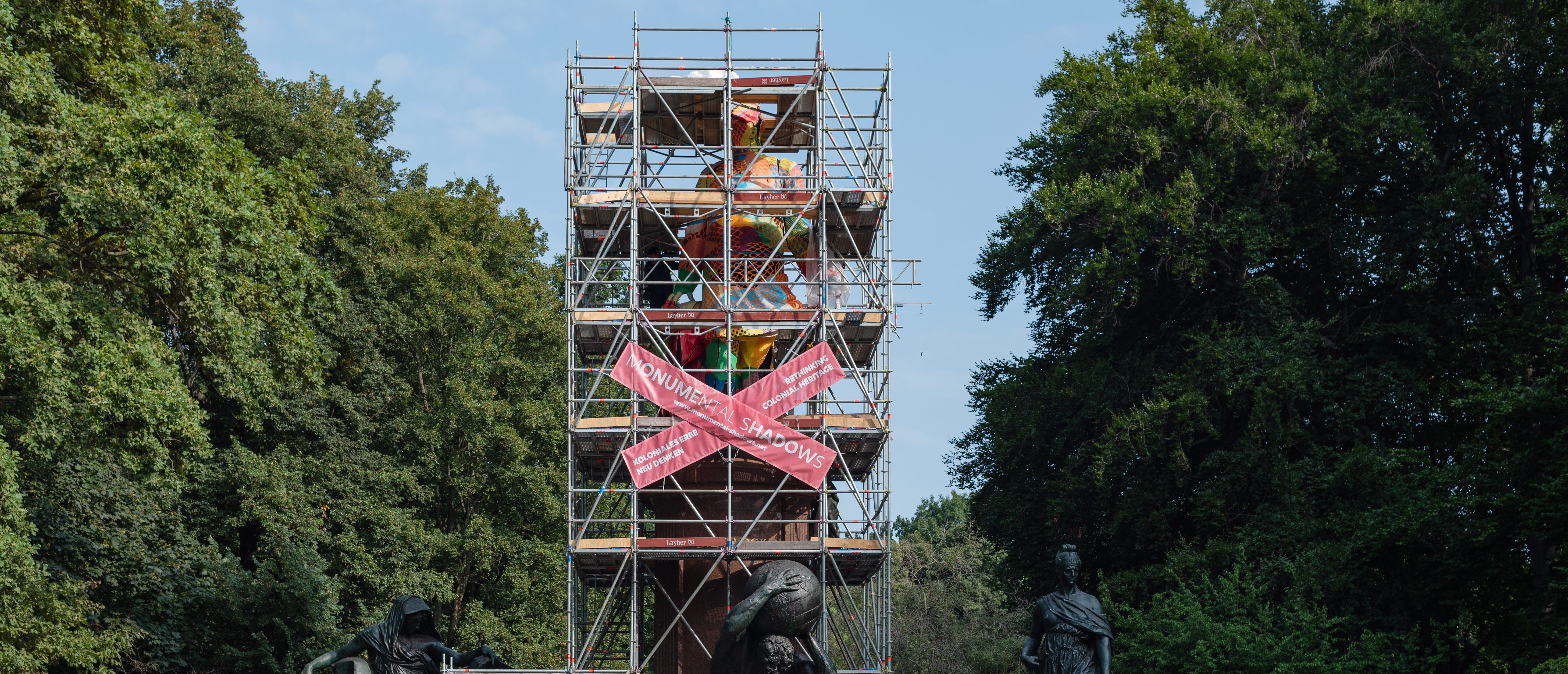 Das Bismarck-Denkmal in Berlin Tiergarten. Ein Baugerüst hüllt die Statue ein. Auf dem Gerüst sind zwei rosa Banner überkreuzt gehängt, auf denen in weißer Schrift "Monumental Shadows" steht. Die Statue ist zum Teil mit buntem Pappmaché eingehüllt.