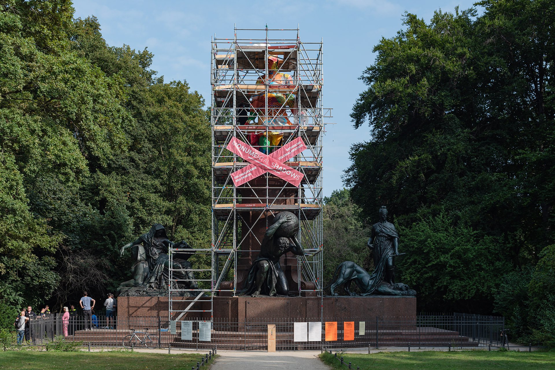 Das Bismarck Denkmal in Berlin Tiergarten. Ein Baugerüst hüllt die Statue ein.  Auf dem Gerüst sind zwei rosa Banner überkreuzt gehängt auf denen in weißer Schrift "Monumental Shadows" steht. Die Statue ist zum Teil mit buntem Pappmaché eingehüllt.
