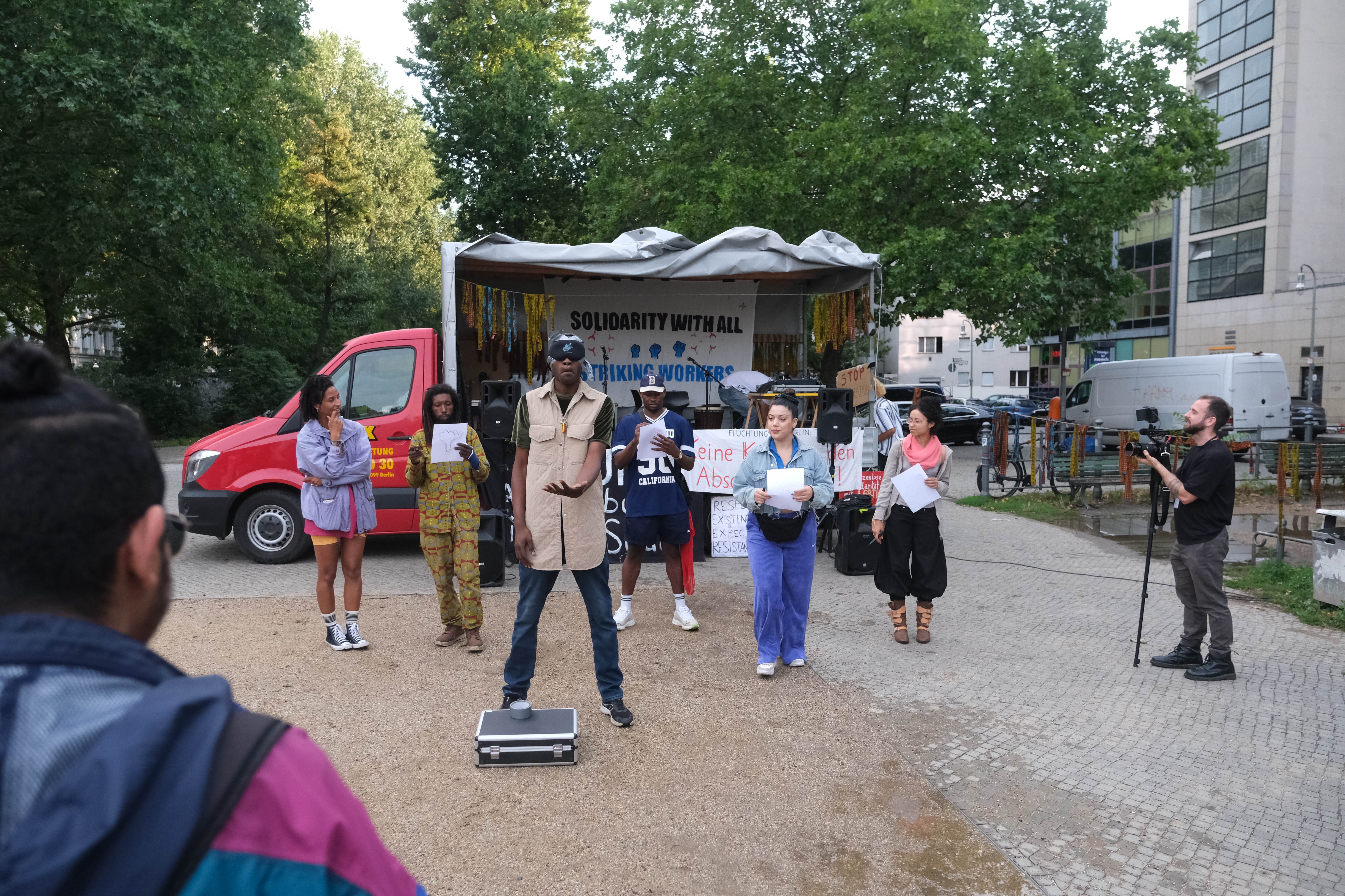 Auf dem Oranienplatz steht ein Auto mit einem offenen Laster. Davor stehen sechs Personen, die etwas performen.