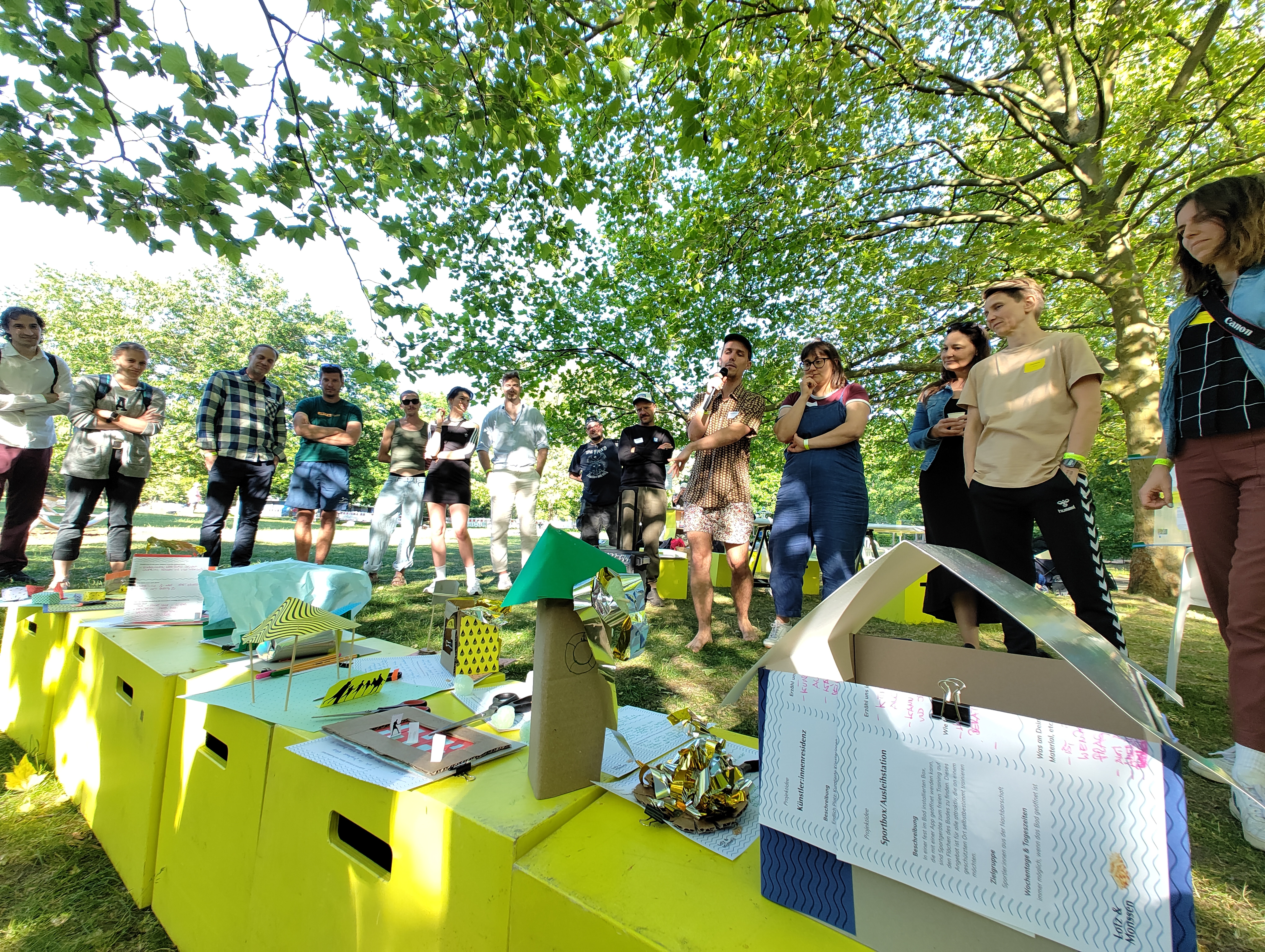 Im Vordergrund sind Sitzblöcke in einem Park aufgebaut, auf denen aus Pappe gebastelte Häuser stehen. Im Hintergrund steht eine Menschengruppe, welche auf die gebastelten Häuser blickt. Eine Person hat ein Mikro in der Hand und spricht.