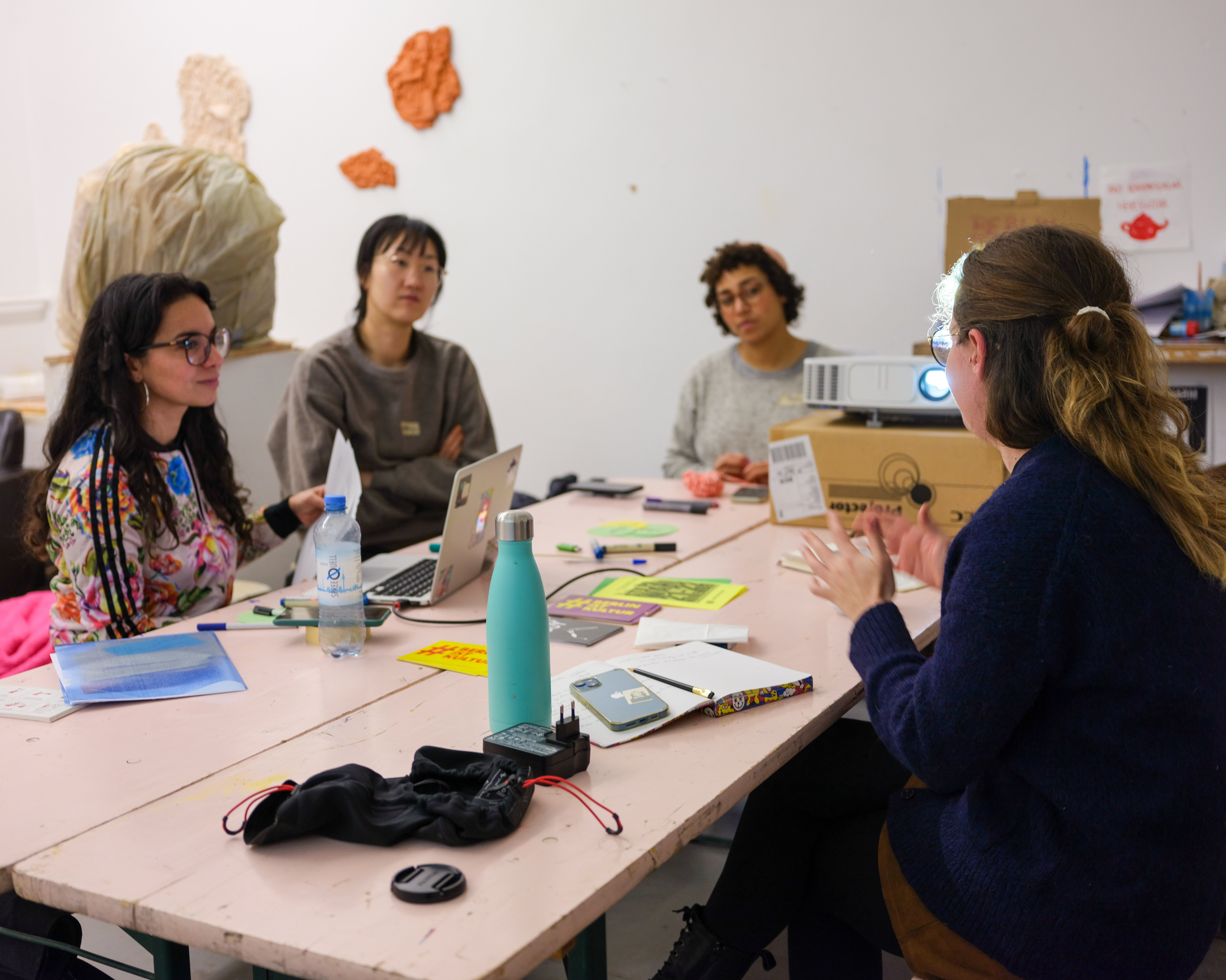 Vier Frauen sitzen um einen Tisch herum. Laptops, Notebooks, Wasserflaschen und andere Materialien sind auf dem Tisch ausgebreitet. Auf einem Pappkarton ist ein Projektor aufgestellt.  Im Hintergrund sind Wandkunst und Materialien zu sehen.