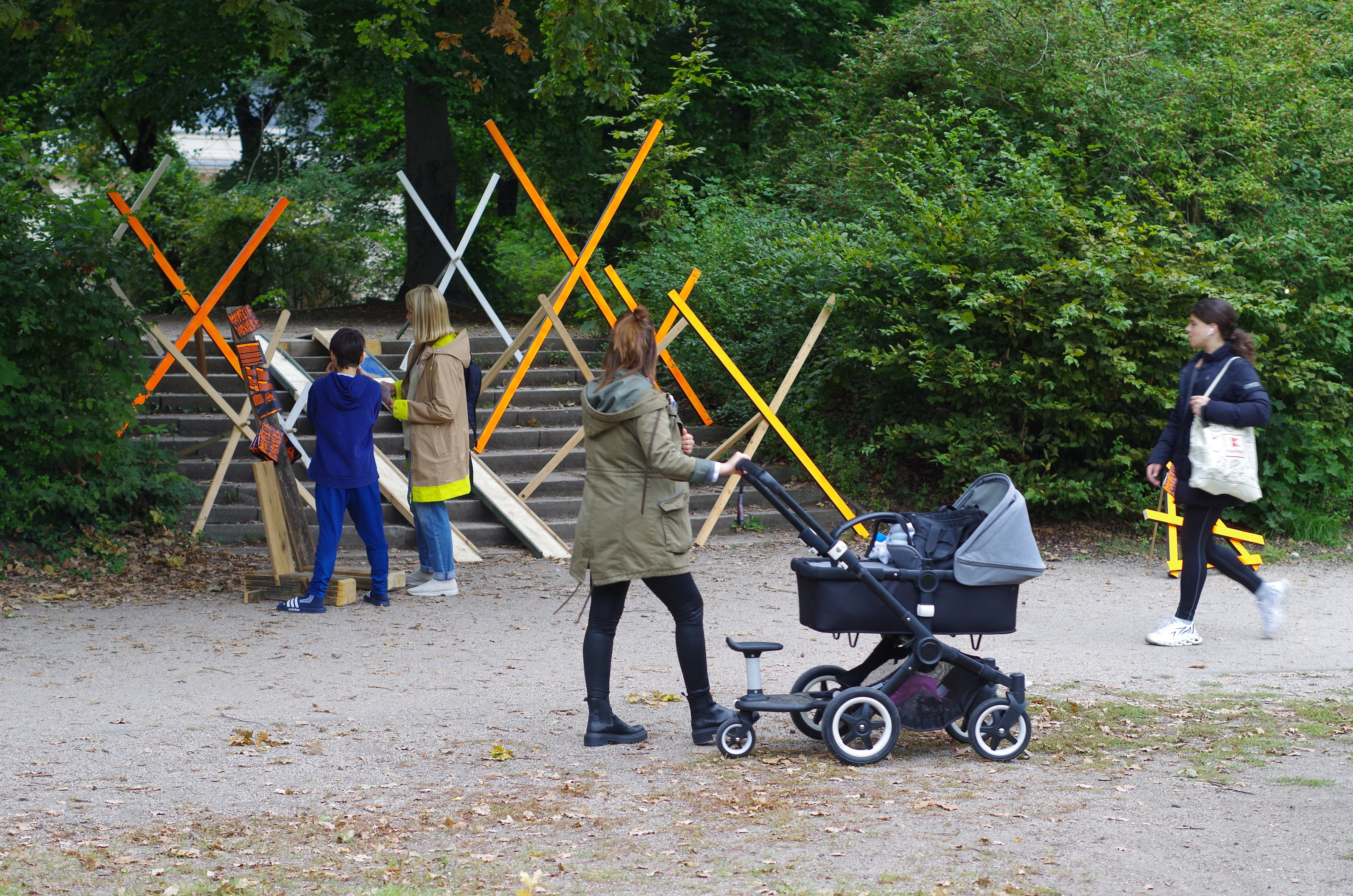 Eine breite Treppe im Bürgerpark Pankow, auf deren Stufen insgesamt acht bunte, als X aufgestellte Holzlatten verteilt sind. In der Mitte der Treppe führt eine provisorische Rampe die Treppe rauf. Eine Person mit Kinderwagen läuft vorbei.