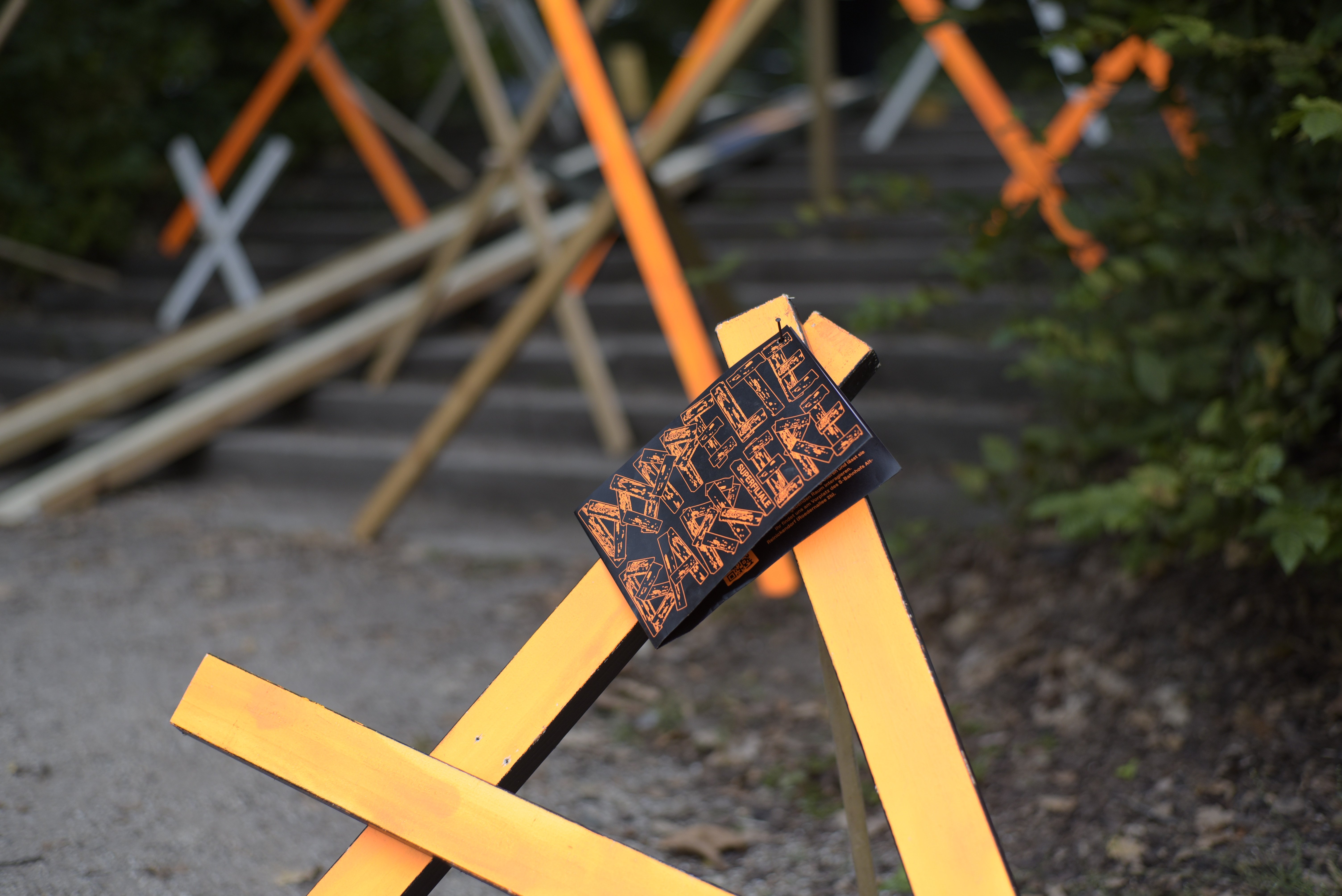 Auf dem Bild sind im Ausschnitt zwei orange bemalten Holzlatten zu sehen, die spitz aufeinander zu laufen. An der Spitze ist ein Flyer mit der Aufschrift „Doppelte Barriere“ genagelt.