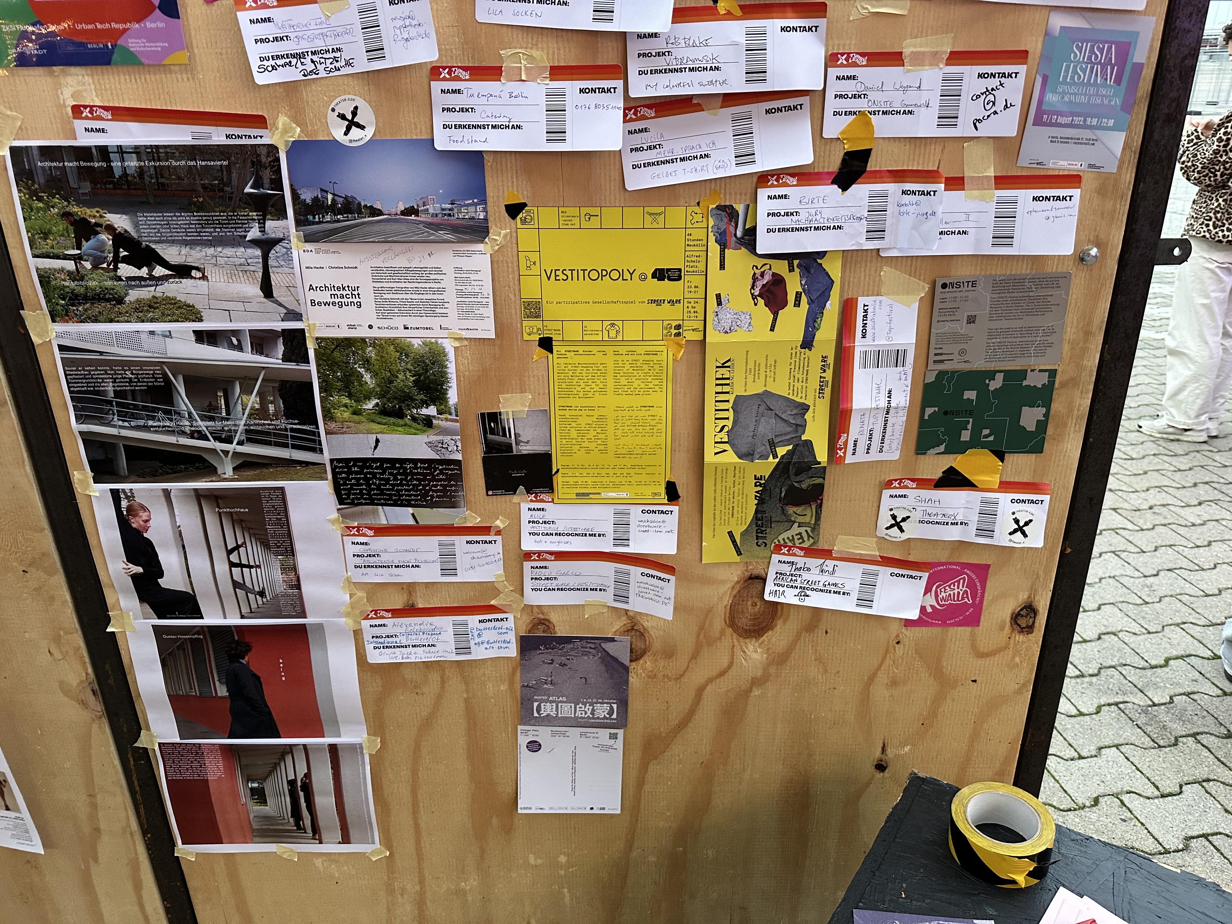 Eine Holzwand ist mit Fotos und Beschreibungen von Projekten beklebt. Dazwischen hängen Karten im Design von Flugzeug-Boardingpässen auf denen zusätzlich Informationen zu den Projekten stehen.
