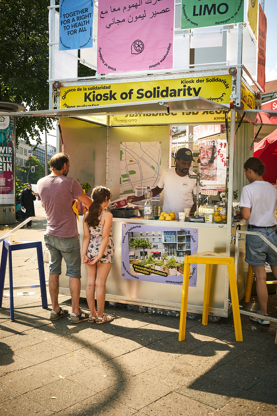 Das Baugerüst des Kiosk ist zu sehen auf dem Bürgersteig einer großen Straße zu sehen. In dem offenen Ladenbereich steht ein Mann und bereitet eine Limonade vor. Vor dem Kiosk stehen einer älterer Mann und ein jüngeres Mädchen.