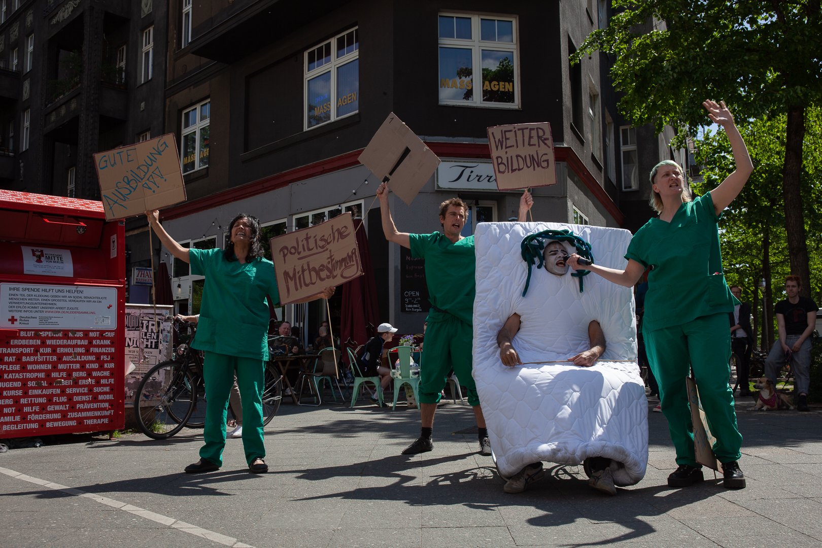 4 Personen stehen auf einem Platz. Drei sind in grüne Arbeitskleidung von Pflegepersonal in Krankenhäusern gekleidet. Sie tragen Schilder. Eine Person steckt in einer Matratze und nur der Kopf, die Füße und die Arme schauen aus der Matratze heraus.