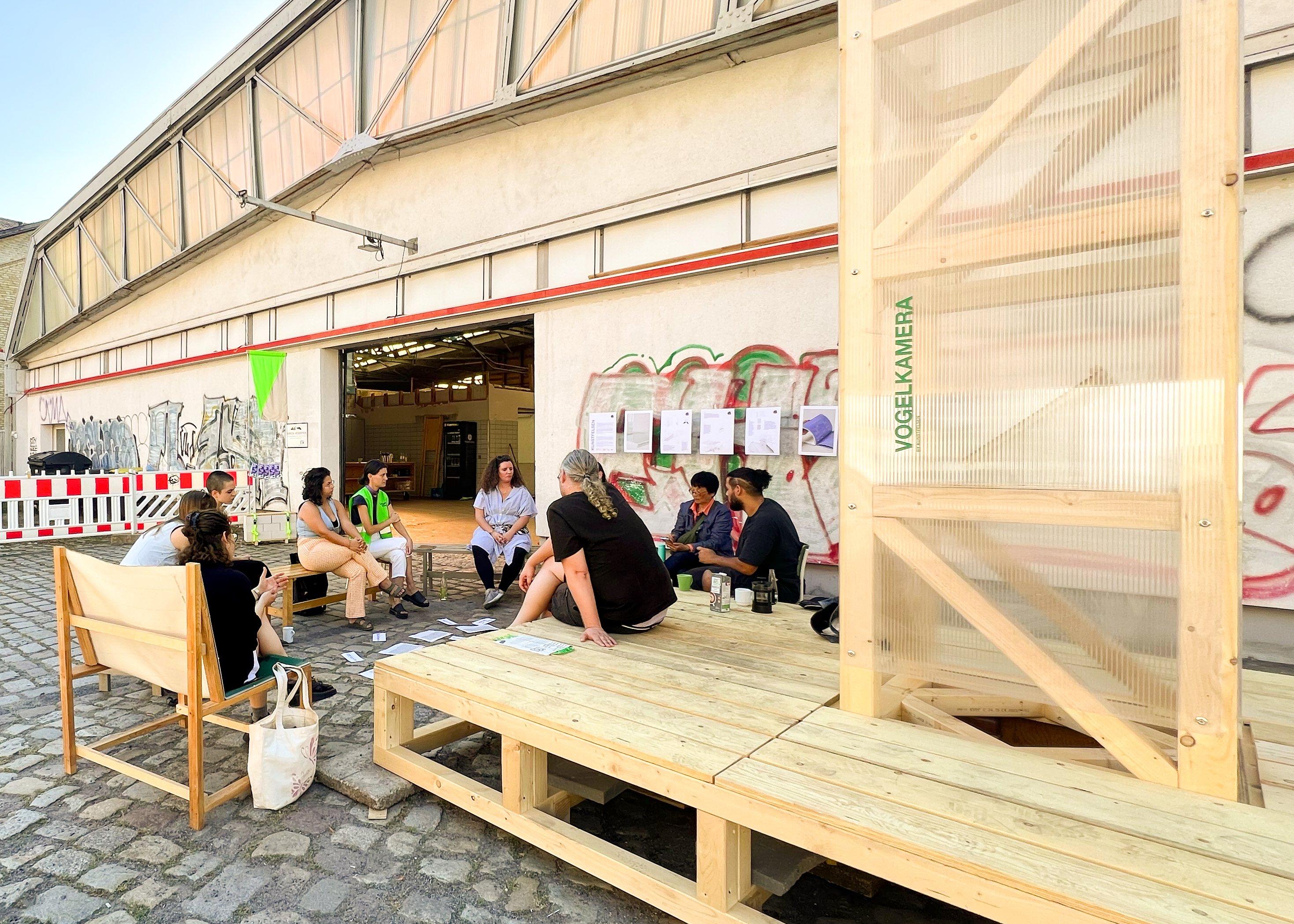Eine Gruppe an Menschen sitzt in einem Kreis vor einer großen Halle.