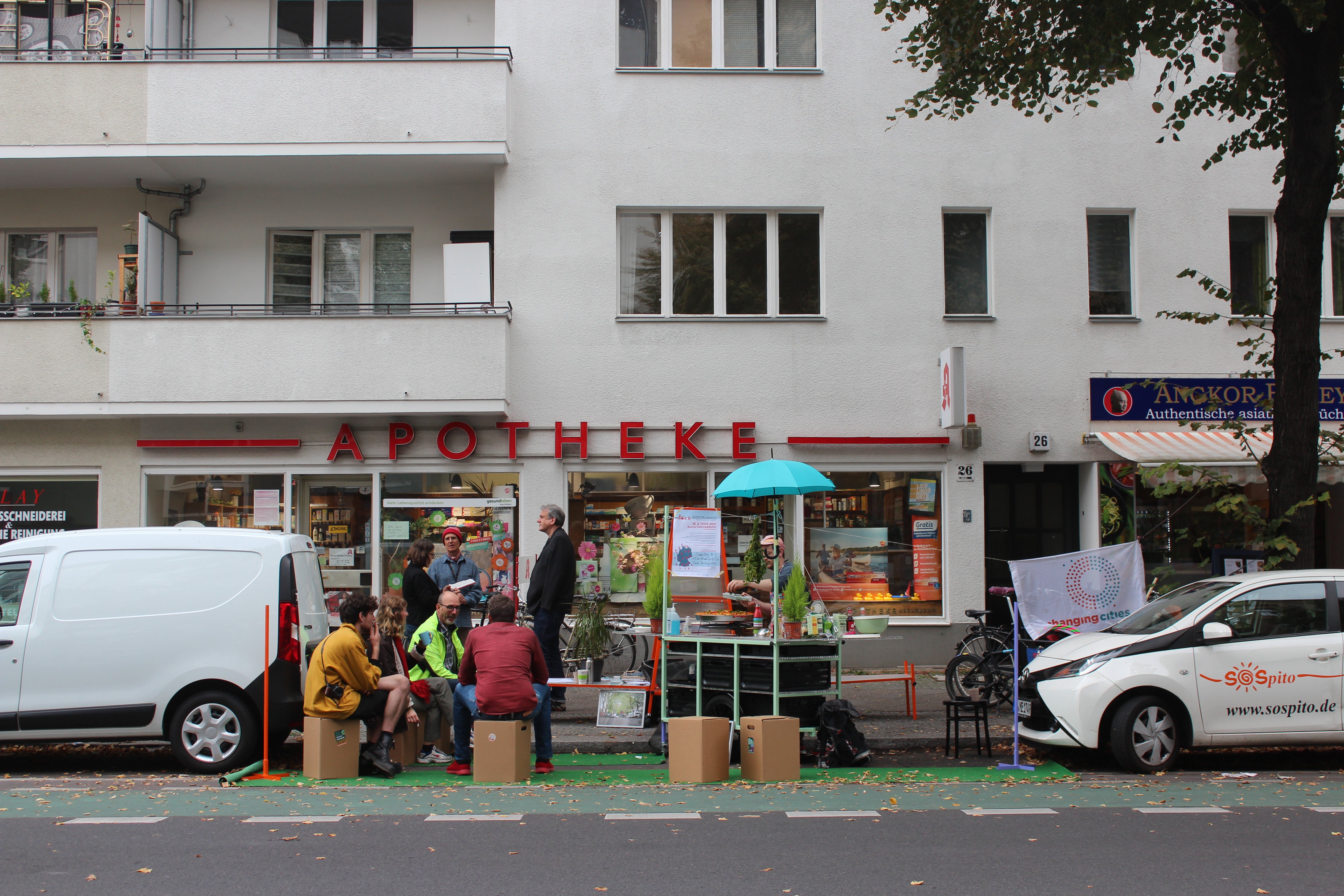 Vor einer Apotheke blockieren die Künstler*innen einen Auto-Parkplatz. Sie haben Papp-Hocker aufgebaut. Einige Menschen sitzen darauf. Rechts daneben steht ein Fahrrad-Anhänger, umgebaut zu einer mobilen Küche. Eine Person schenkt Essen aus.
