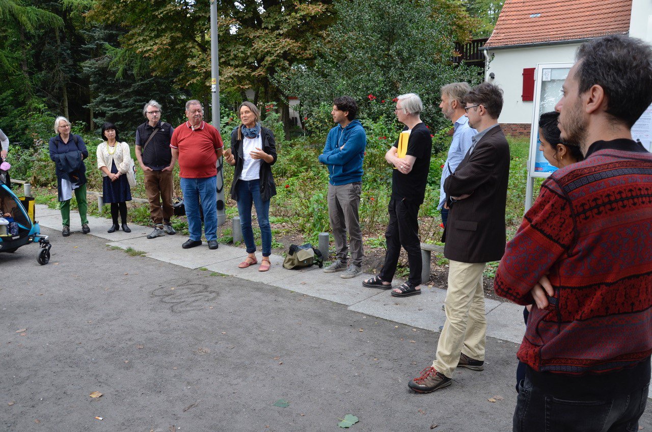 11 Personen stehen im Park um den Schäfersee. Eine Person mit langen grauen Haaren erklärt etwas. Hinter der Gruppe ist ein verwilderter Garten und ein älteres Haus zu sehen. Am linken Bildrand beginnt der Wald oder Park.
