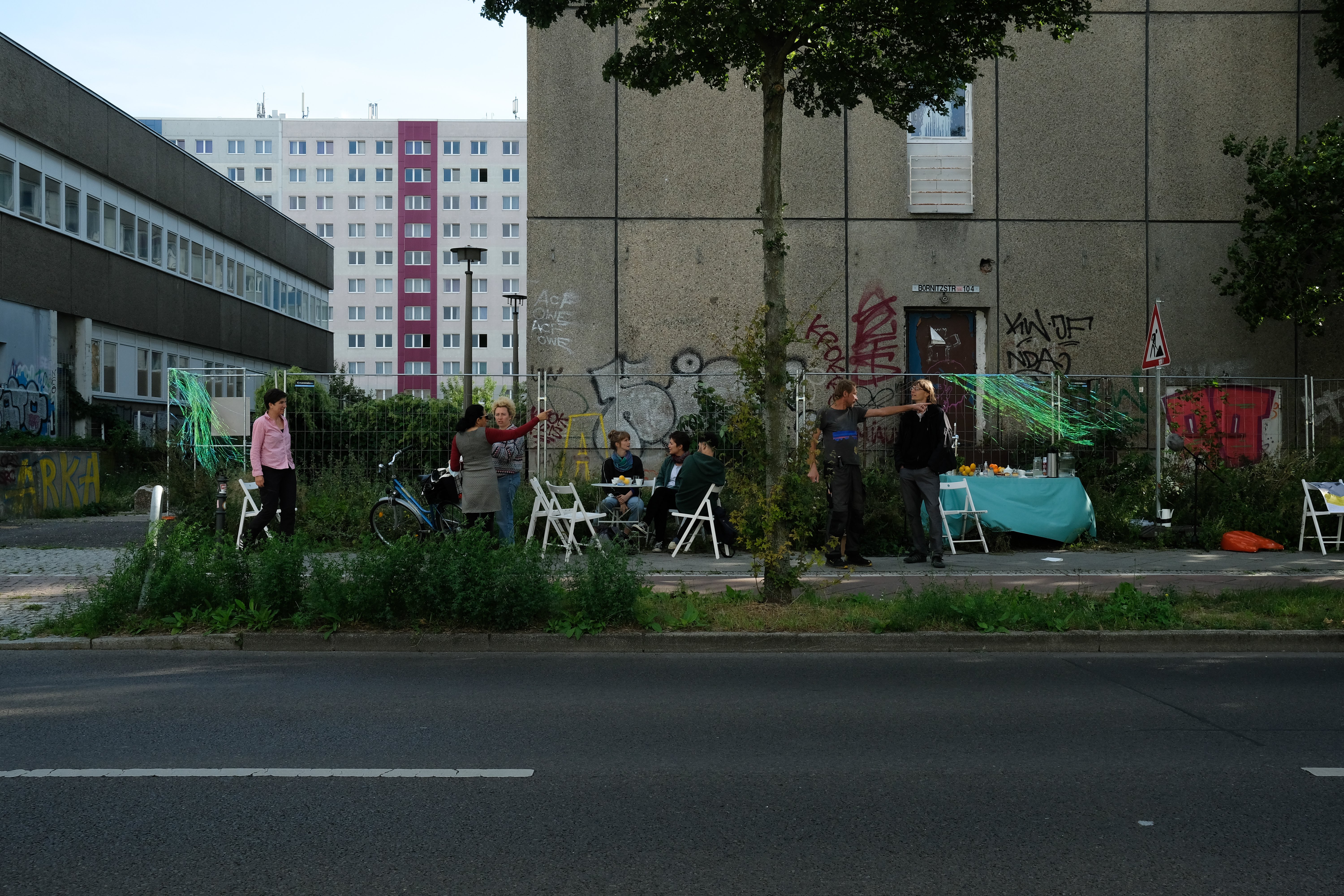 Das Bild zeigt den Ausschnitt zeigt in einer Totalen eine Straße in Berlin-Lichtenberg. Menschen sitzen mit Klappstühlen und Tischen auf dem Bürgersteig. Auf den Tischen sind Kaffeetassen, Obst und Getränke zu sehen. Im Hintergrund stehen verschiedene P