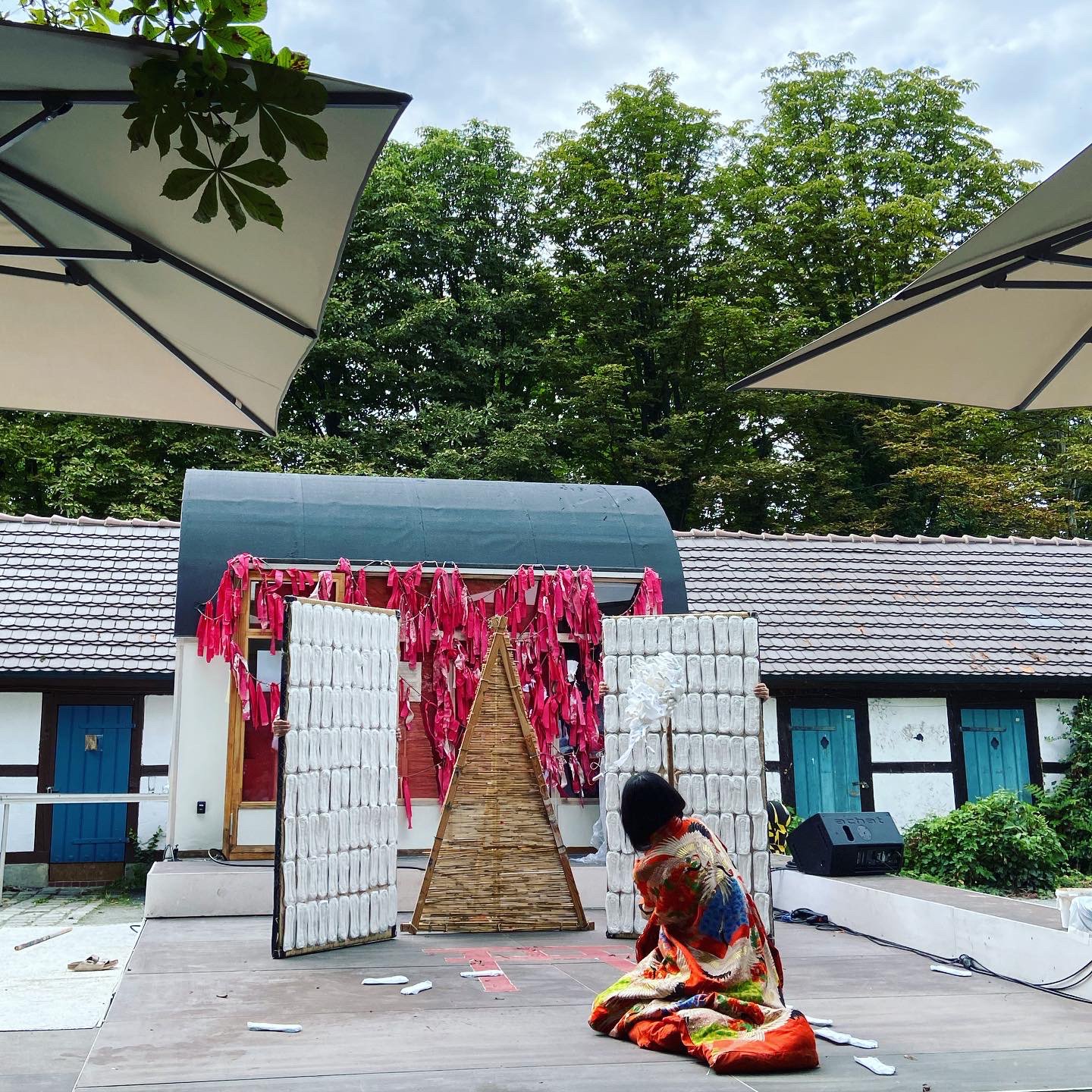 Das Bild zeigt eine Bühnenszene aus der Performance 398_SAKUYA., die von der japanischen Mythologie inspiriert ist. Das Bild zeigt von hinten eine Person in einem roten Gewand mit Applikationen die auf der Bühne kniet.