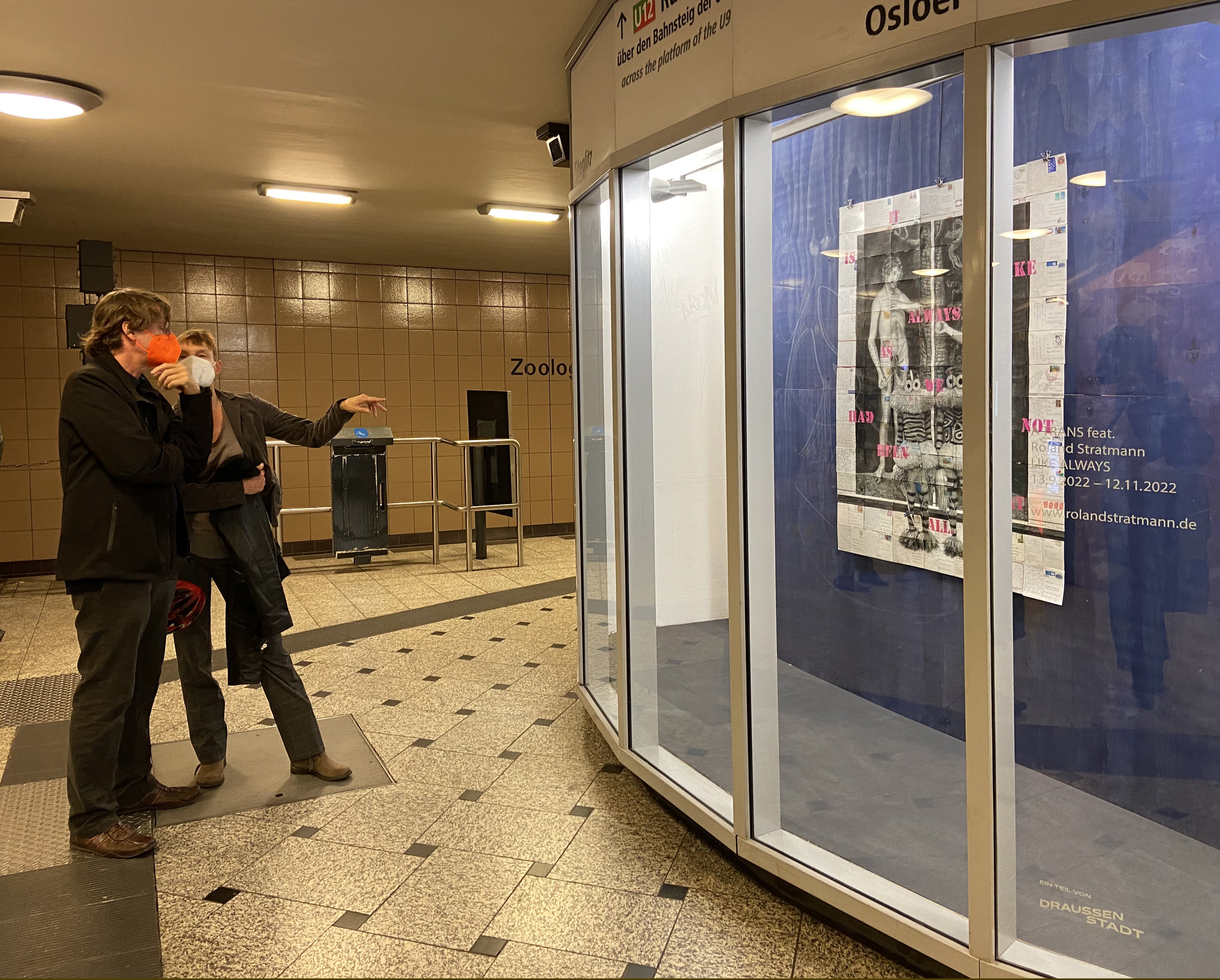 Zwei Menschen stehen vor der Vitrine im U Bahnhof Zoologischer Garten. Sie schauen sich die Collage an.