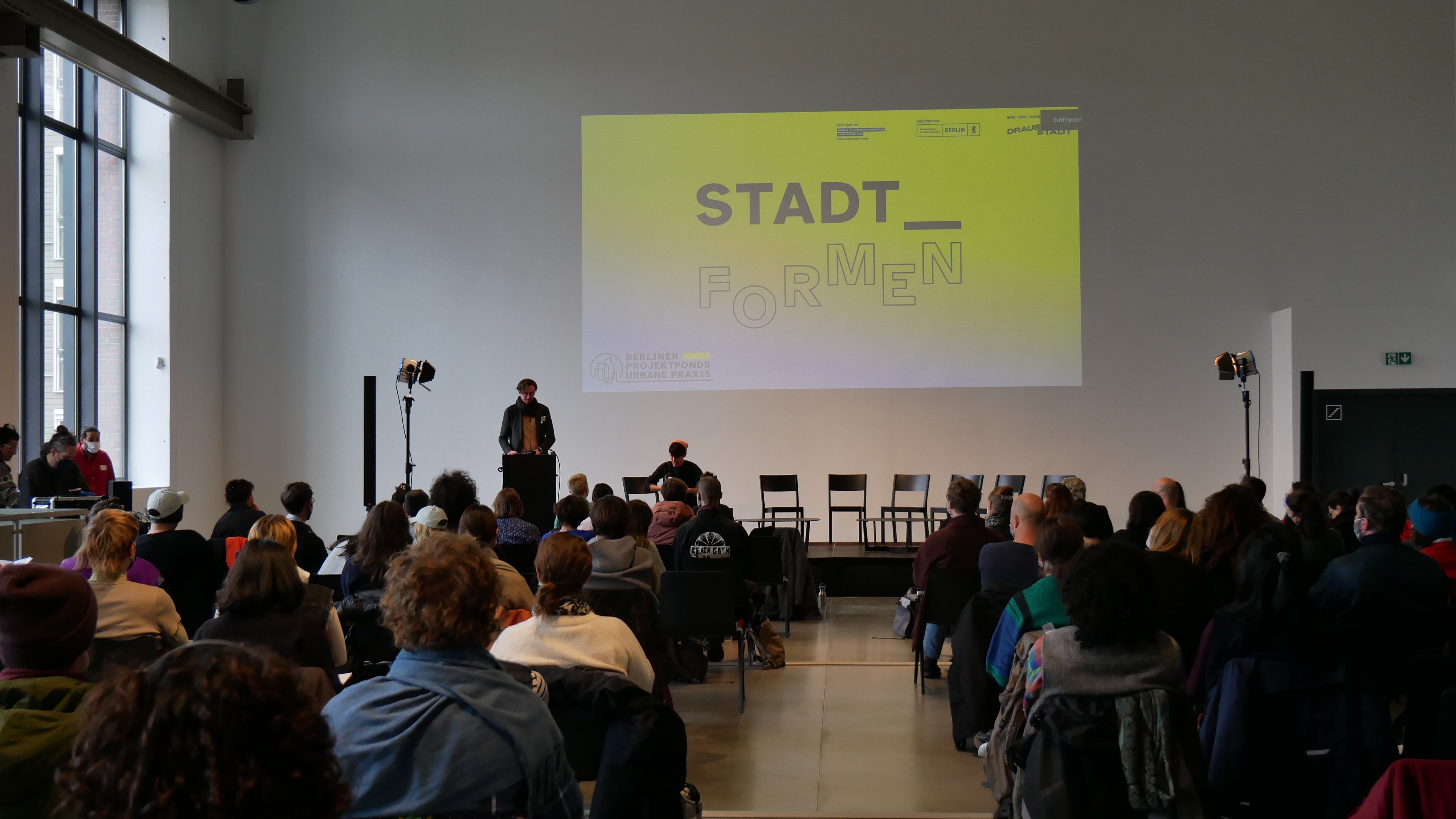 Ein großer Konferenzsaal. Viele Menschen sitzen auf Stühlen vor einer Bühne. Auf der Bühne spricht ein Mensch an einem Podium. Im Hintergrund ist ein Bild an die Wand gebeamt. Dort steht:Stadt_formen