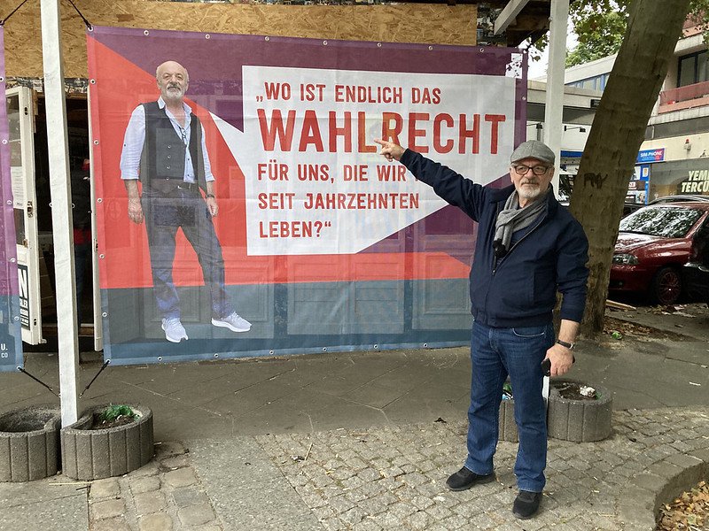 Eine Person Ende 50 mit Schirmmütze zeigt auf ein Plakat hinter sich. Das Plakat am Gecekondu. Auf dem Plakat ist dieselbe Person zu sehen mit der Aufschrift: Wo ist endlich das Wahlrecht für uns die wir seit Jahrzehnten hier leben?