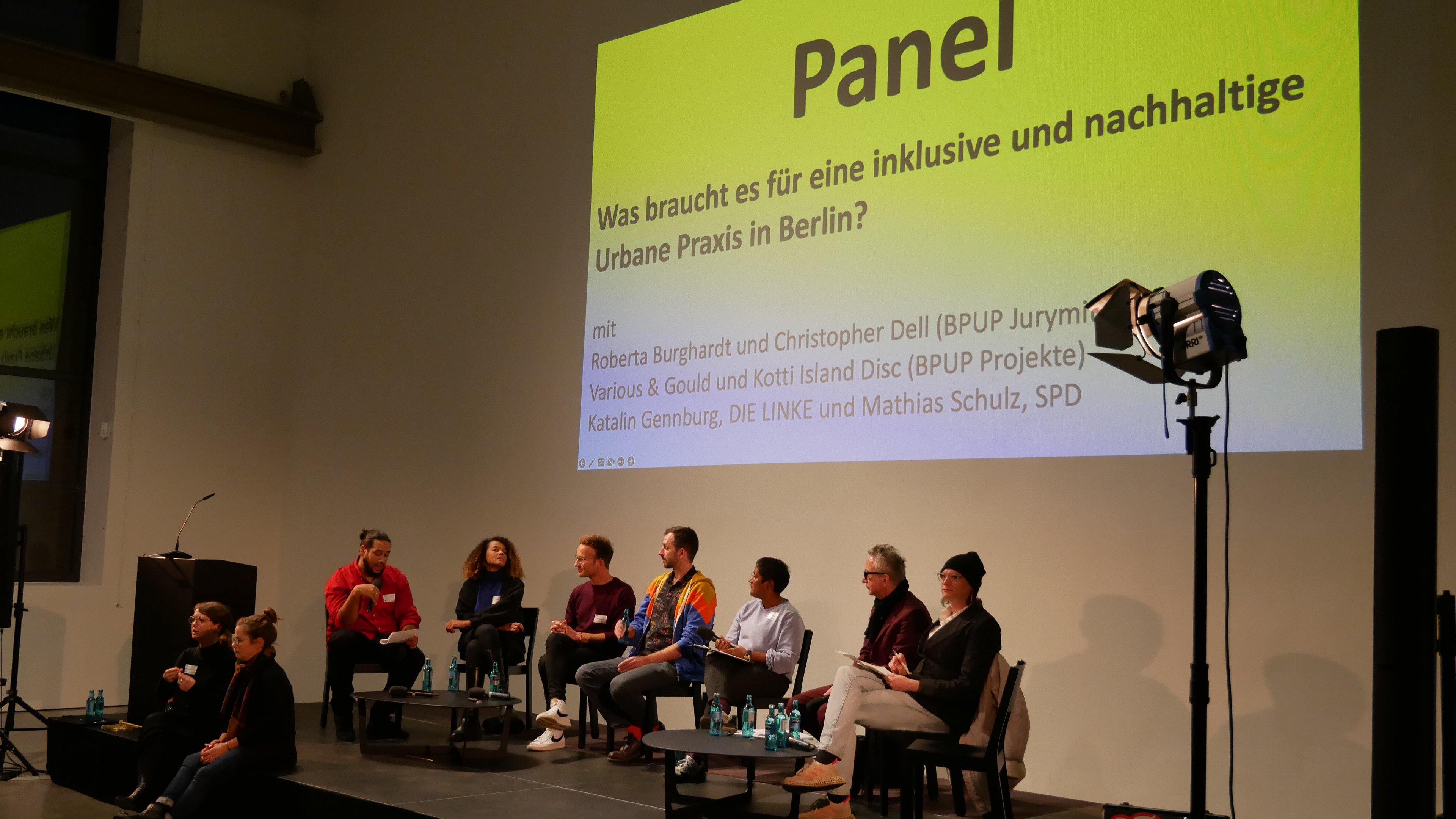 Sieben Personen sitzen auf einer Bühne. Über ihren Köpfen ist eine Folie an die Wand gebeamt. Darauf steht: Panel. Was braucht es für eine inklusive und nachhaltige Urbane Praxis in Berlin?.