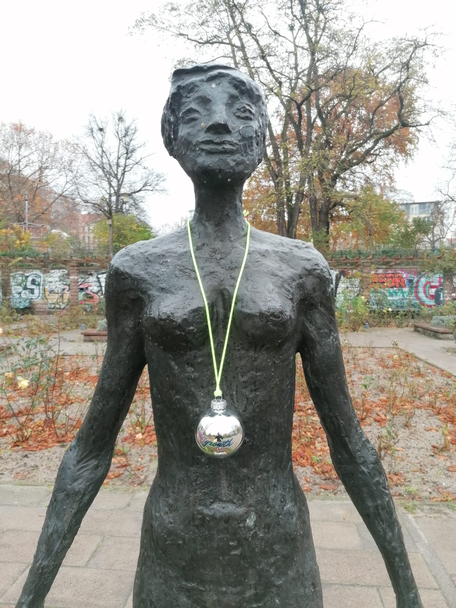 Eine schmale Frauen-Statue in einem Park in Berlin-Mitte mit offenen Armen und stolzem Blick nach oben. Eine der Weihnachtskugeln der Kunstaktion hängt um ihren Hals.
