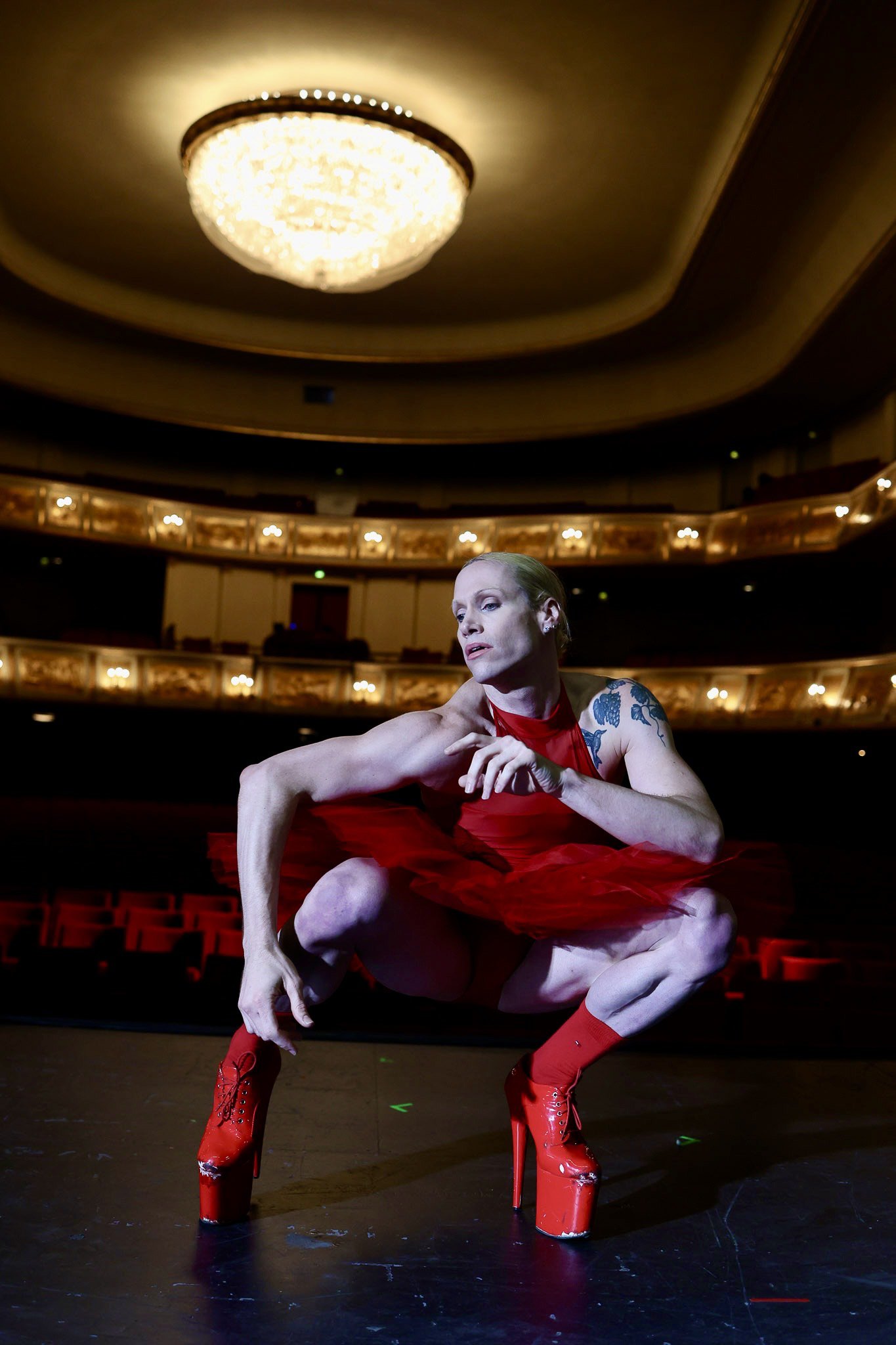 Ein Foto von Lucio Vidal. Die Person hat ein rotes Ballettkleid an und hohe rote Schuhe. Die Person hockt auf einer Theaterbühne.