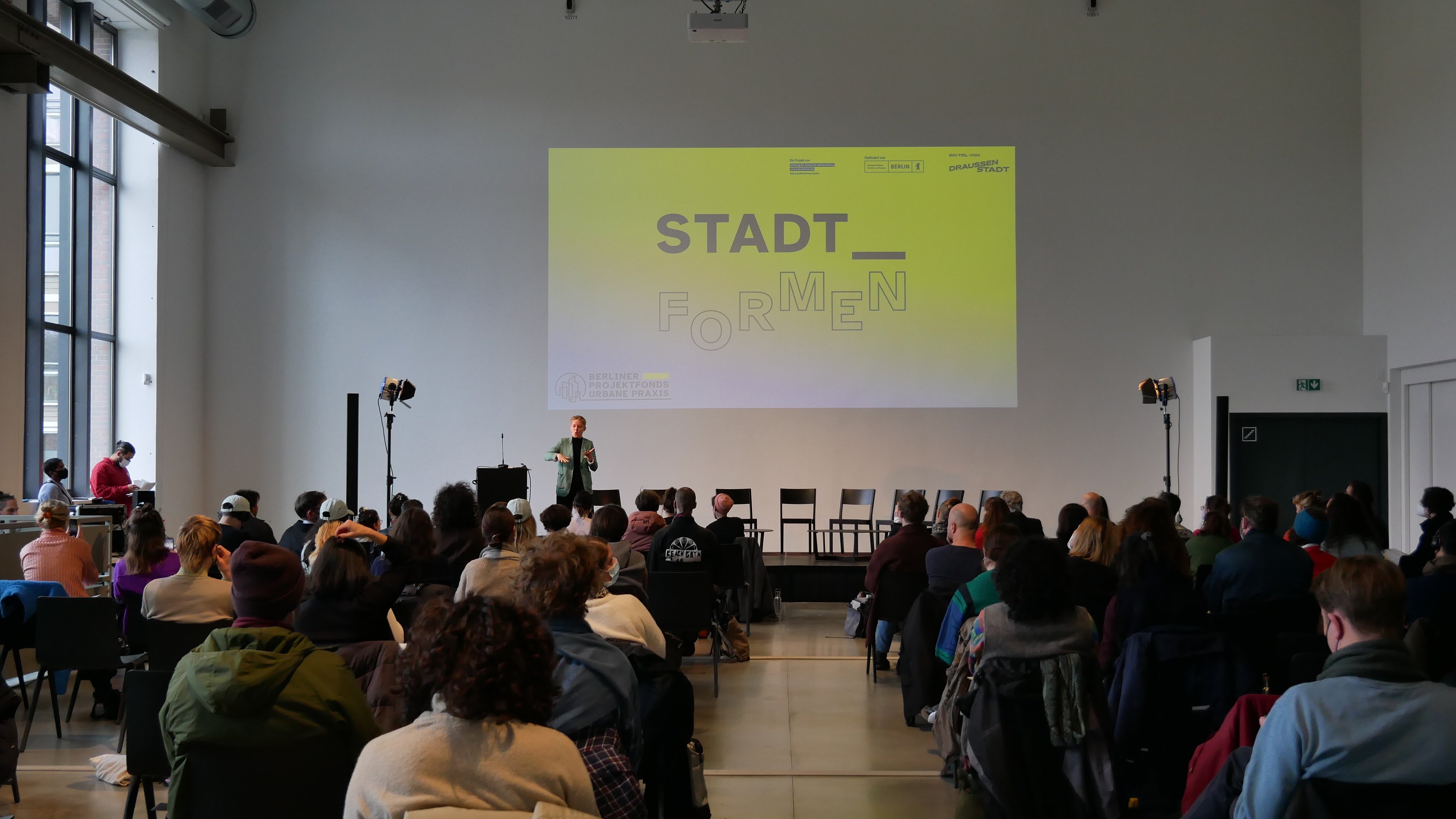 Vor einer Bühne sitzen viele Menschen auf Stühlen und blicken auf eine Gebärdende Person, die auf der Bühne steht. Über der Bühne ist eine Folie an die Wand gebeamt auf der steht: Stadt_formen. Darunter ist das Logo des Berliner Projektfonds Urbane Praxis