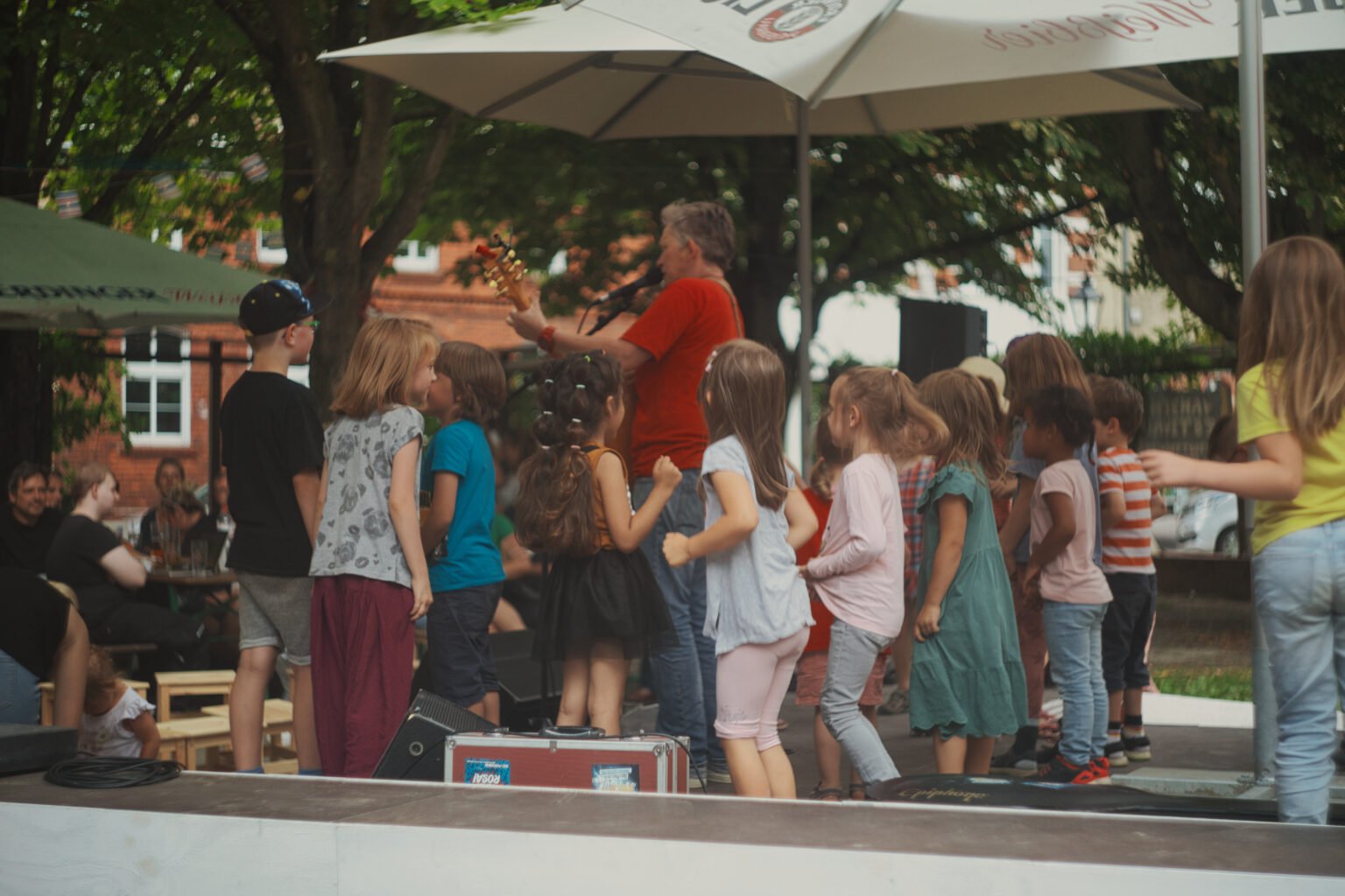 Die Open-Air-Bühne vom Wanzenfestival in Spandau von der Seite. Auf der Bühne steht eine Person mit Gitarre. Drum herum stehen etwa 13 Kinder, vielleicht als Chor.