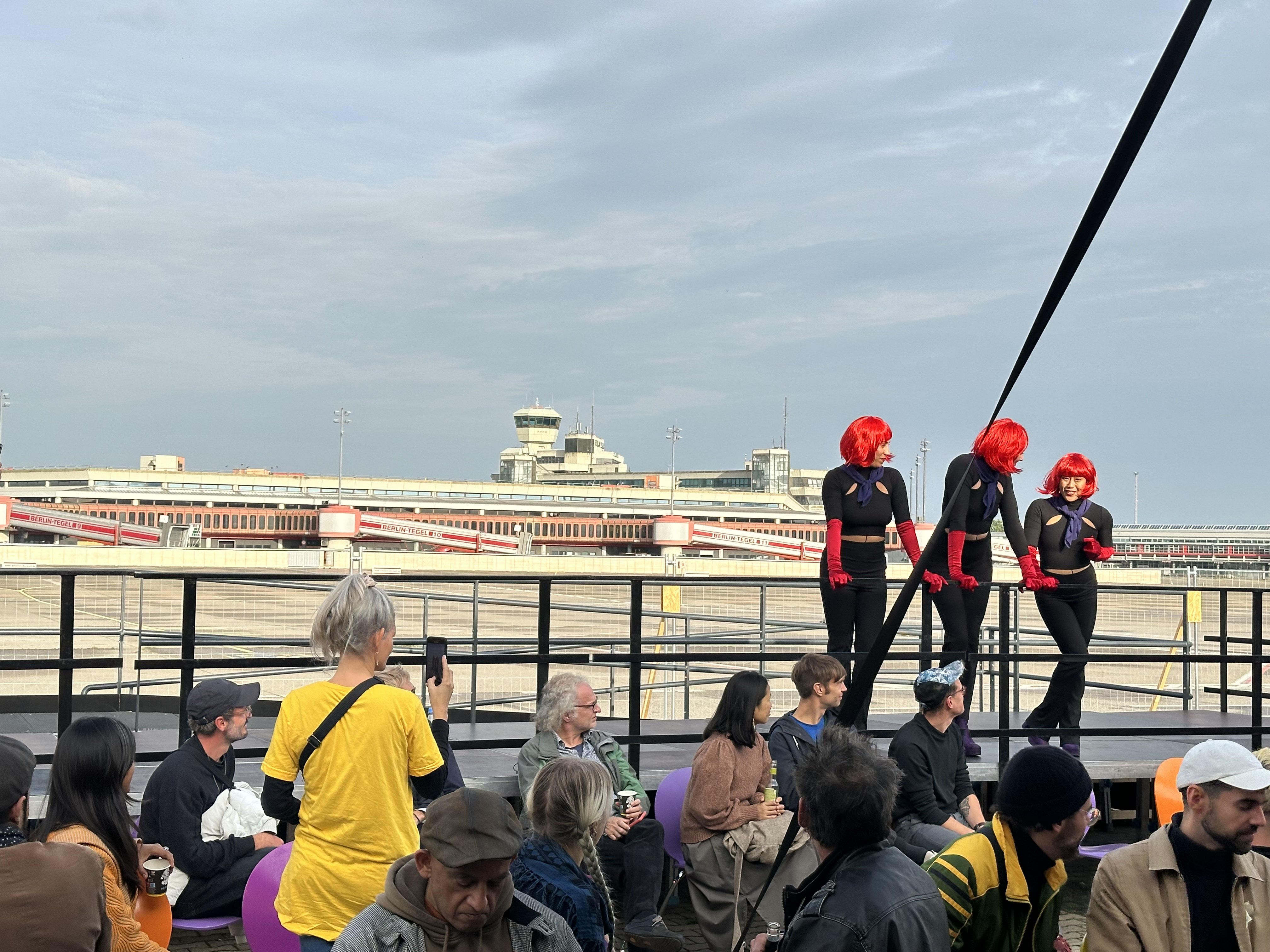 Drei Personen mit roten Perücken und Stewardessoutfits stehen auf einer Tribüne. Im Hintergrund ist der ehemalige Tegel-Flughafen zu sehen. Im Vordergrund sitzen 16 Personen und schauen die drei Personen mit Perücken an.
