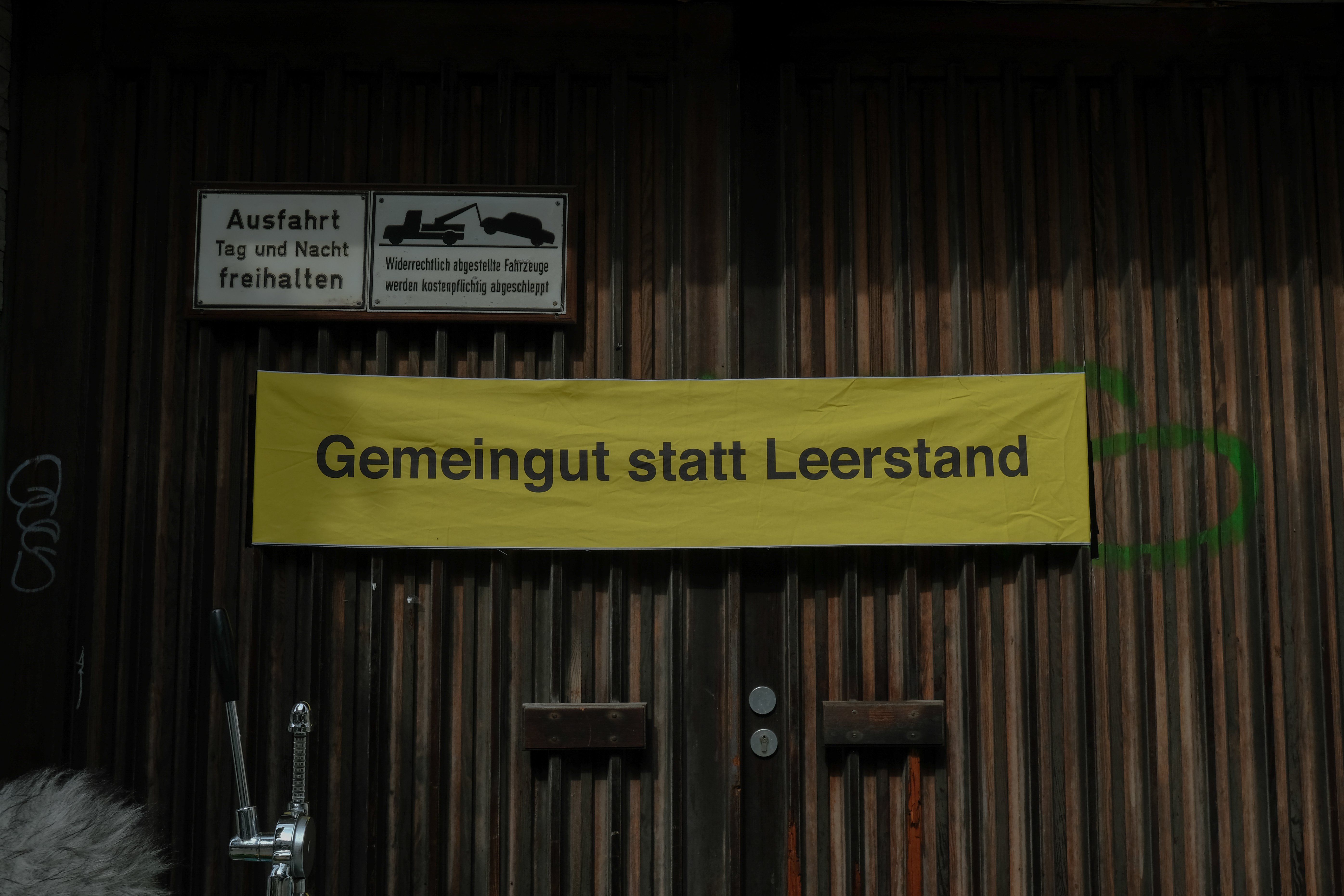 Auf einem hölzernen Garagentor mit den Schildern „Ausfahrt Tag und Nacht freihalten“ und „Widerrechtlich abgestellte Fahrzeuge werden kostenpflichtig abgeschleppt“ hängt ein gelbes Banner. Mit schwarzer Schrift steht darauf „Gemeingut statt Leerstand“.