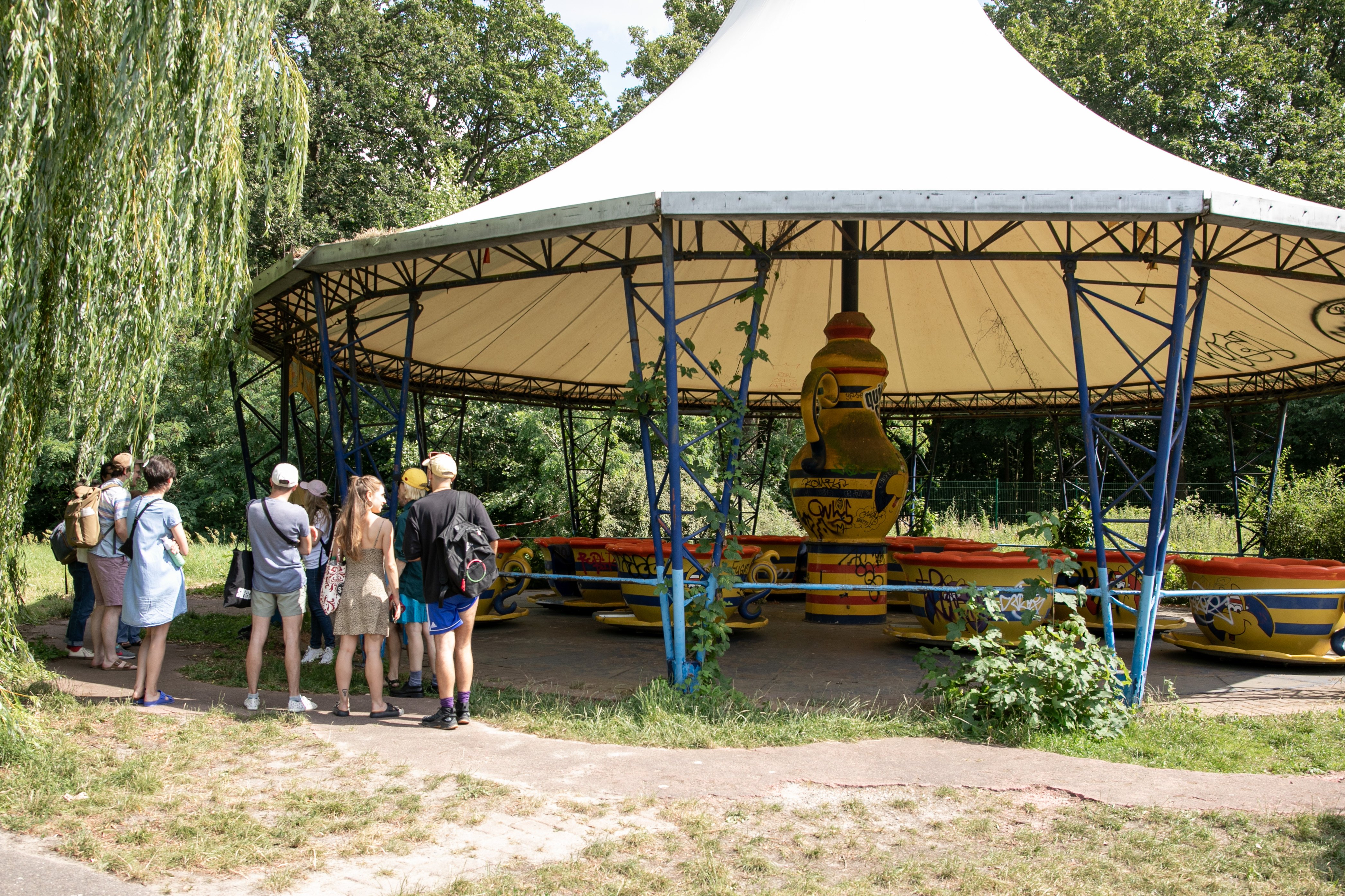 Eine Menschengruppe steht in einem Park vor einem großen spitzen Pavillion. Unter dem Pavillion ist eine Säule mit Verziehrungen.