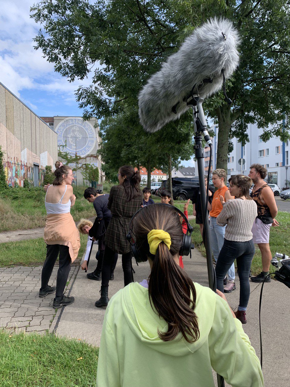 Eine Gruppe an Menschens stehen auf einem Bürgersteig. Im Vordergrund ist ein jüngeres Kind, welches ein Filmmikrophon hält und Kopfhörer trägt.