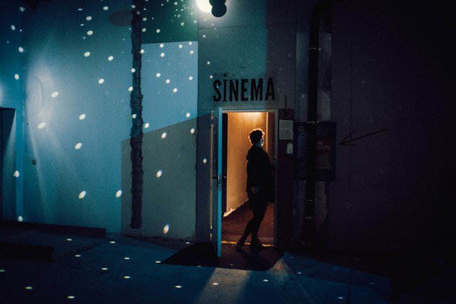 Das Bild ist abends im Foyer des Sinemas aufgenommen. Im Raum sind Lichtprojektionen wie durch eine Diskokugel. Es ist eine Person von hinten zu sehen, die durch eine Tür geht. Über der Tür steht „Sinema“.