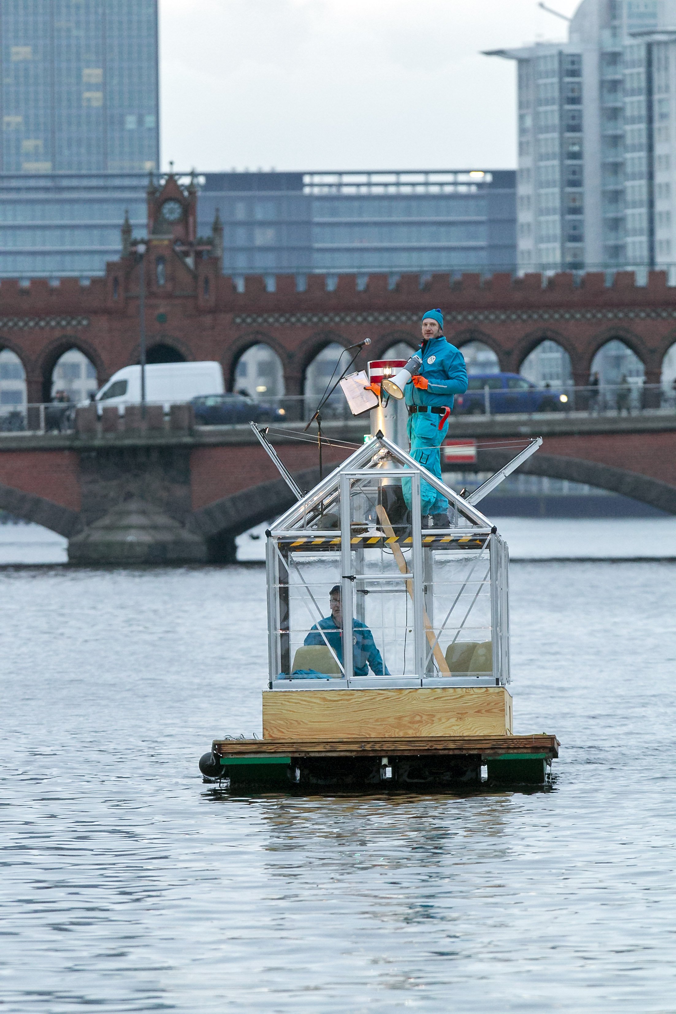 Zwei Personen sind auf dem selbstgebauten Floß auf der Spree vor der Oberbaumbrücke. Eine Person schaut Richtung Kamera aus dem Plexiglas-Dach des Floßes heraus, ein Megaphon in der Hand. Die andere Person steuert das Floß.